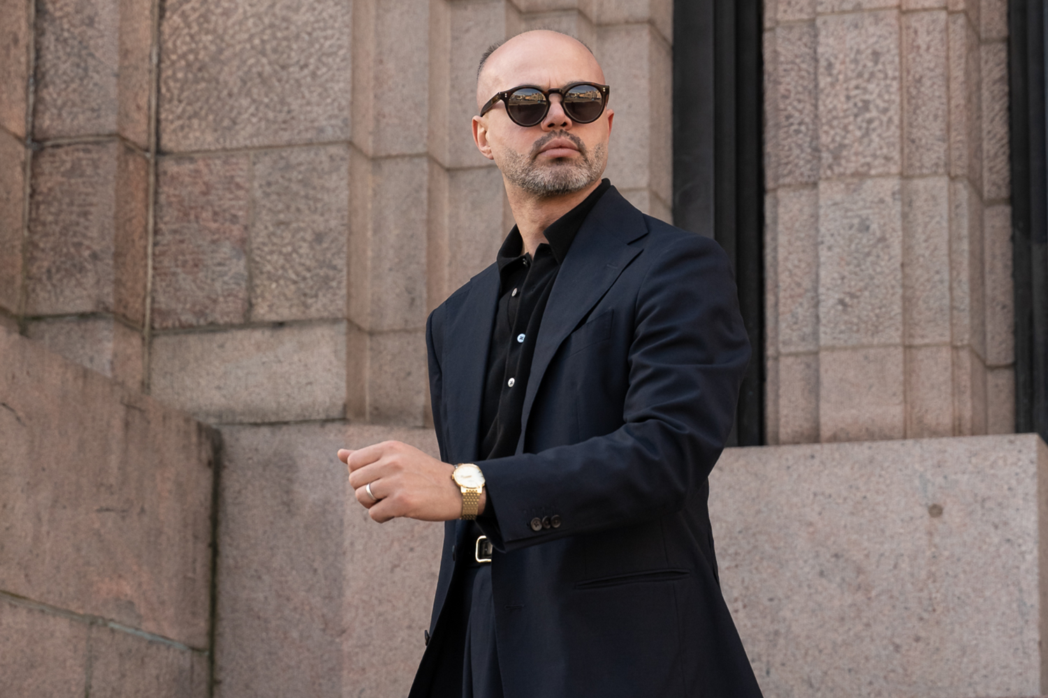 Man with a gold Rotary watch wearing a dark coat and sunglasses standing against a stone building.