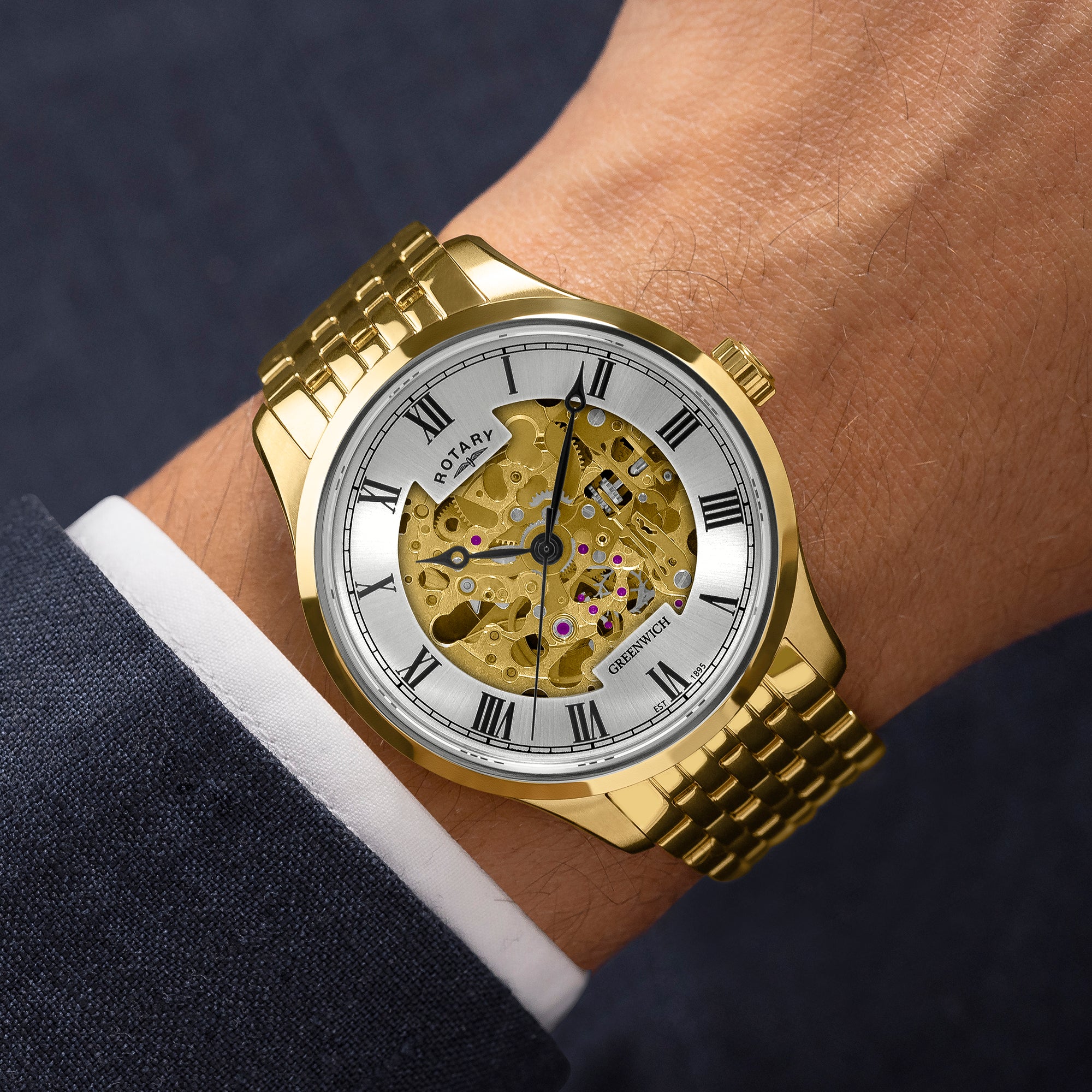 Gold wristwatch with a visible mechanical movement on a wrist, wearing a dark suit and white shirt.
