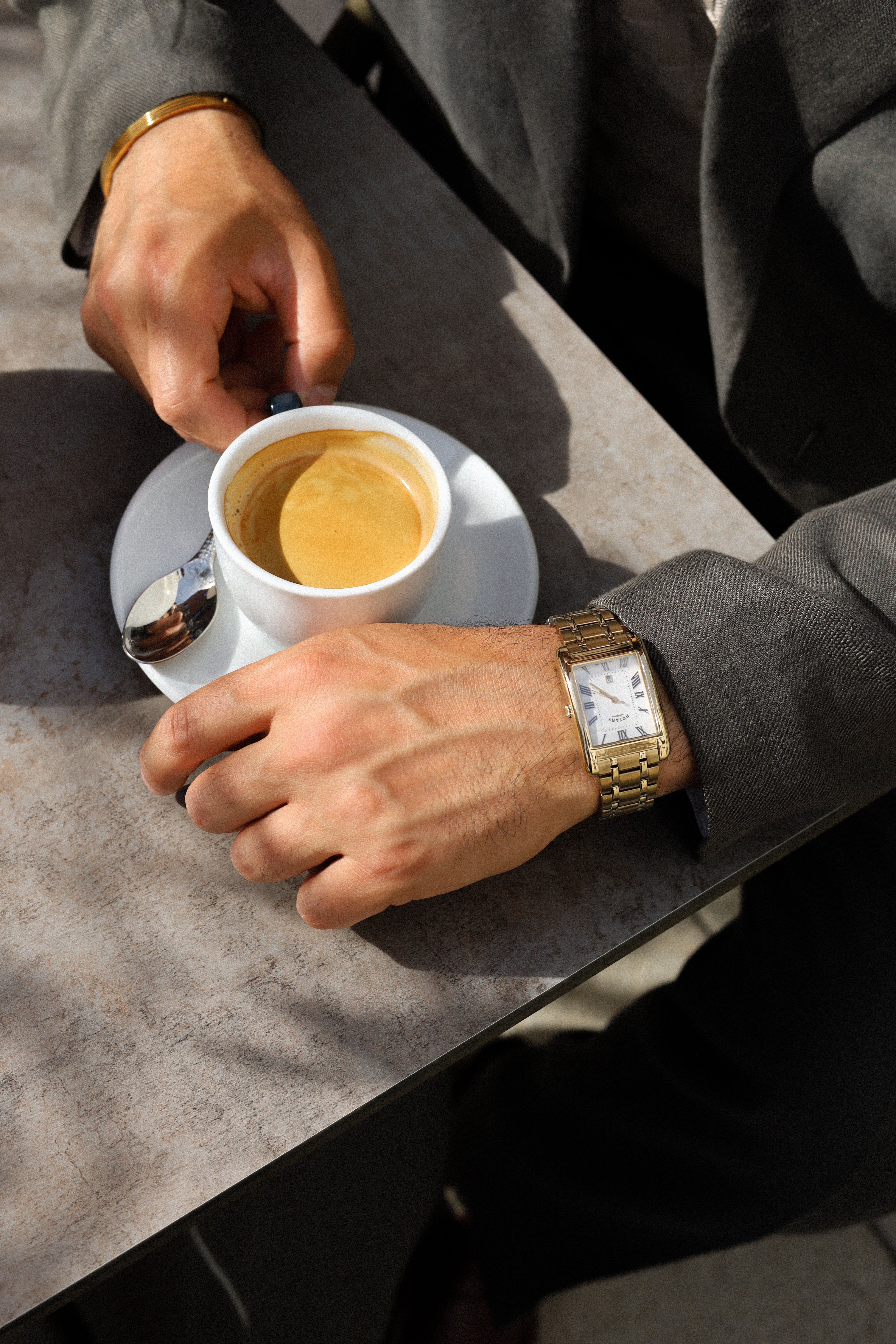 Person holding a small cup of coffee with a saucer on a table wearing a gold Rotary watch.