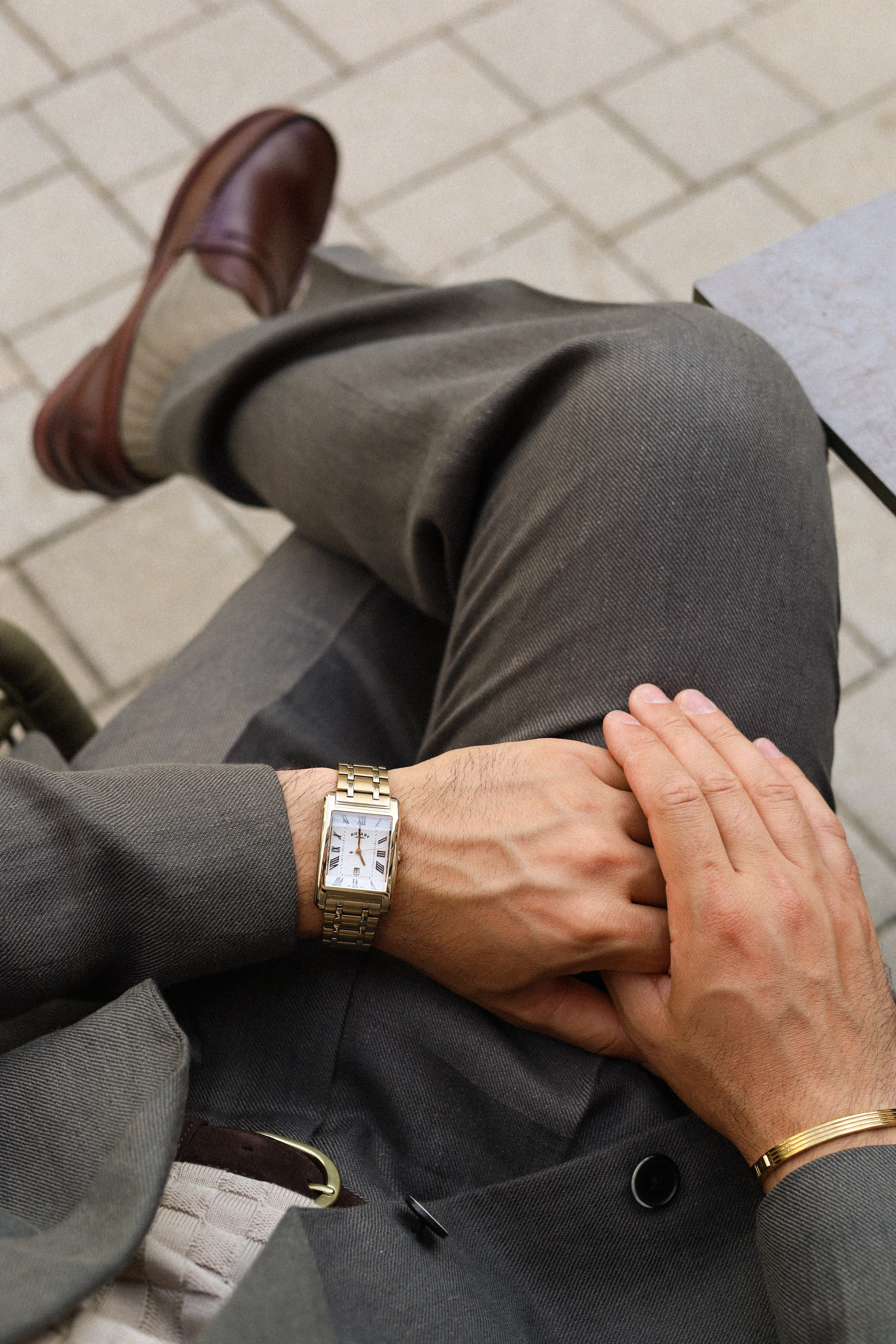 Man wearing a gold Rotary watch and gray suit sitting on a bench.