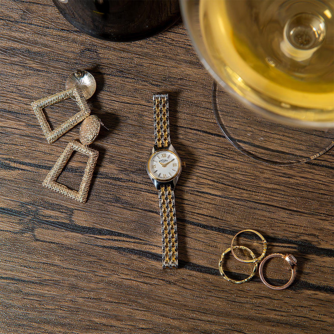 Rotary ladies cocktail watch in two-tone with a white dial. The watch is featured on a wooden table with gold jewellery and a glass of wine