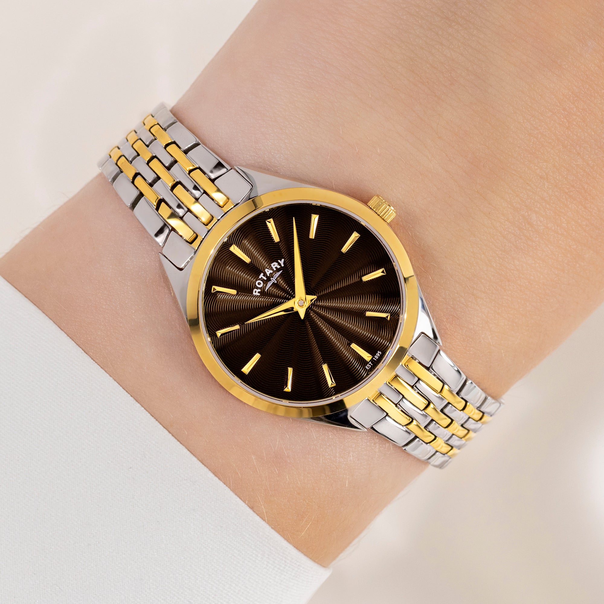 Rotary wristwatch with gold and silver metal strap on a wrist against a neutral background