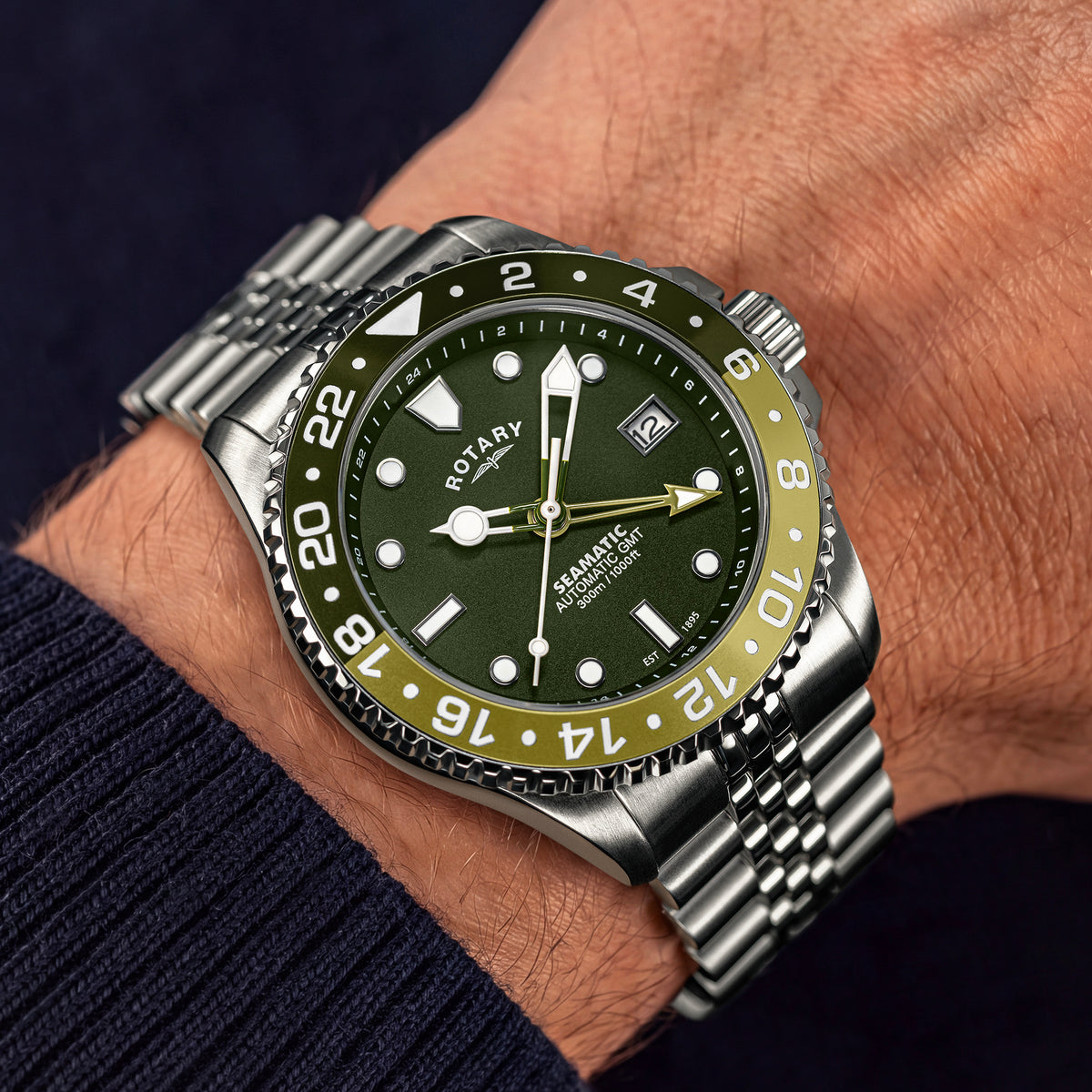 A close-up of a wrist wearing a Rotary Seamatic 300 GMT Automatic watch with a green dial, olive green bezel, and silver stainless steel bracelet.