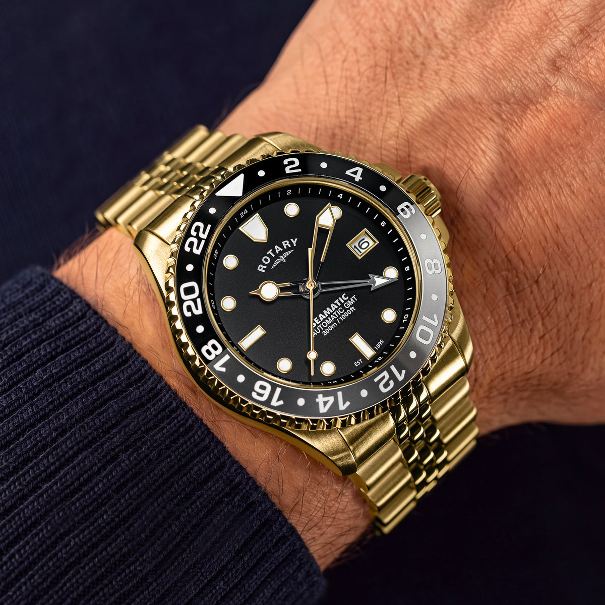 A gold-tone Rotary Seamatic automatic wristwatch with a black dial and black rotating bezel, shown worn on a person's wrist. The watch has gold-tone hour markers, a date window at 3 o'clock, and a matching gold-tone link bracelet.