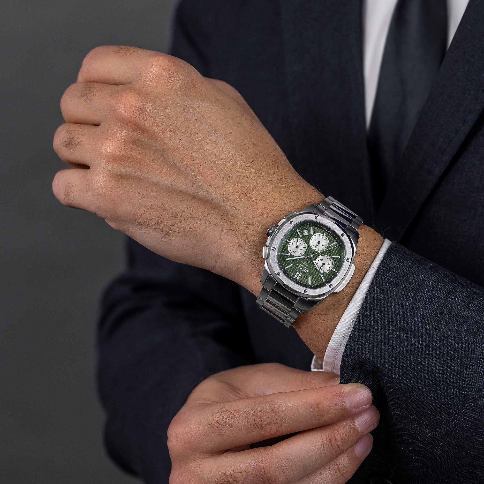 Hand wearing a green Rotary Apex100 Chronograph watch with a dark suit on a gray background