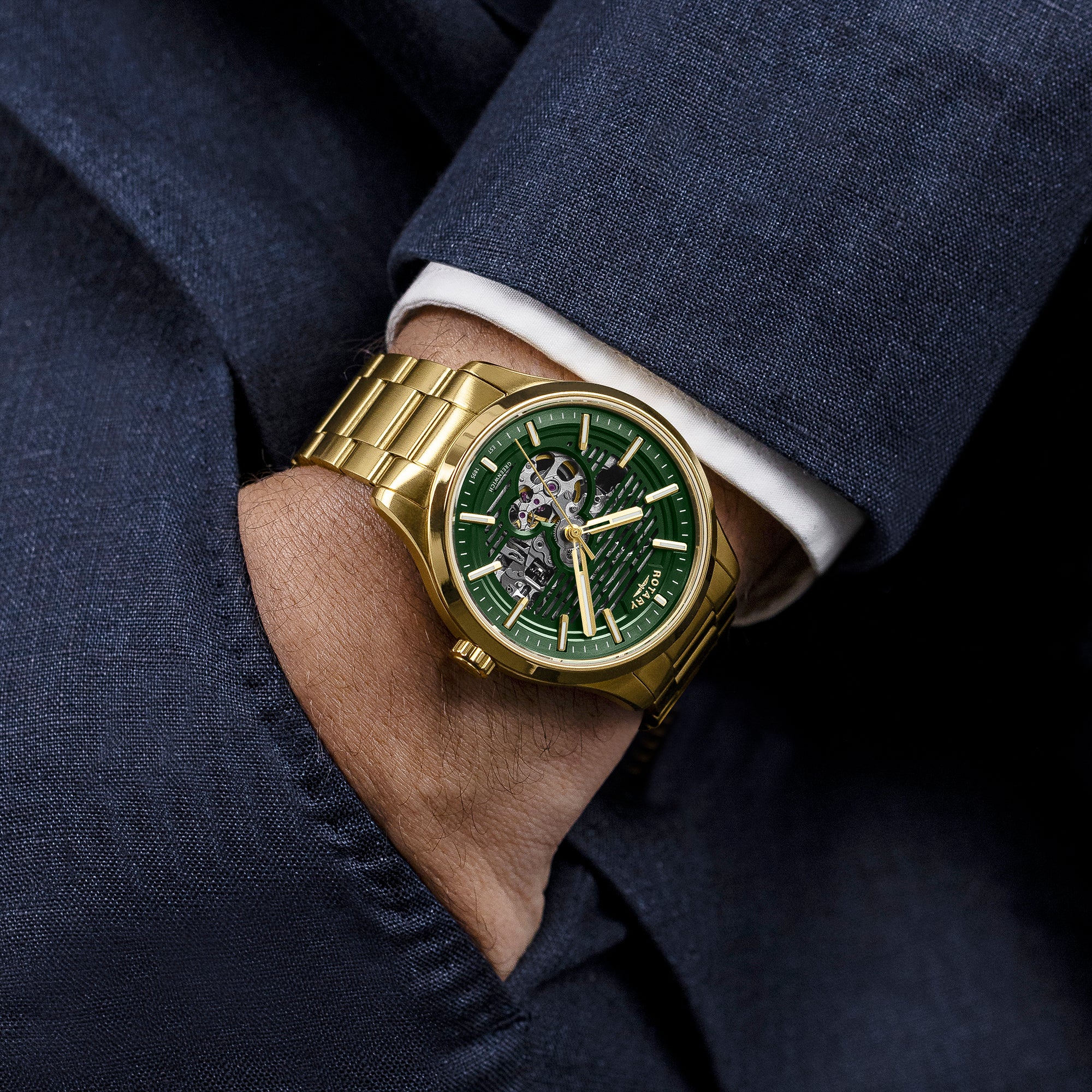 Gold Rotary Greenwich GT1 wristwatch with a green face on a person wearing a dark suit and white shirt.