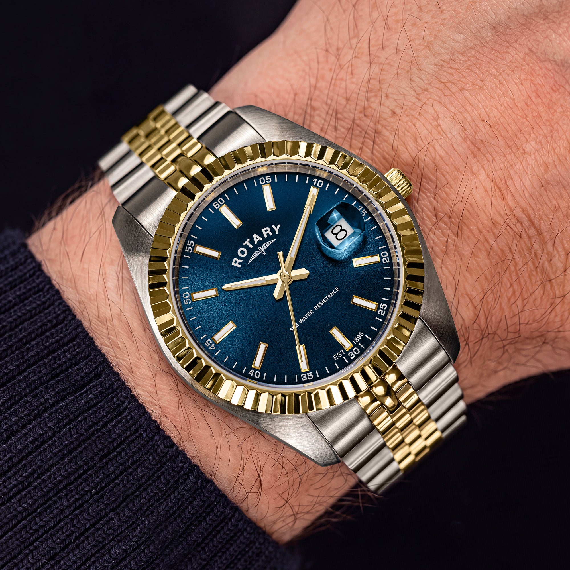 A close-up of a wrist wearing a two-tone metal Rotary watch with a blue dial, gold-tone bezel and hands, and a silver-and-gold link bracelet. The watch brand ROTARY is visible on the dial.
