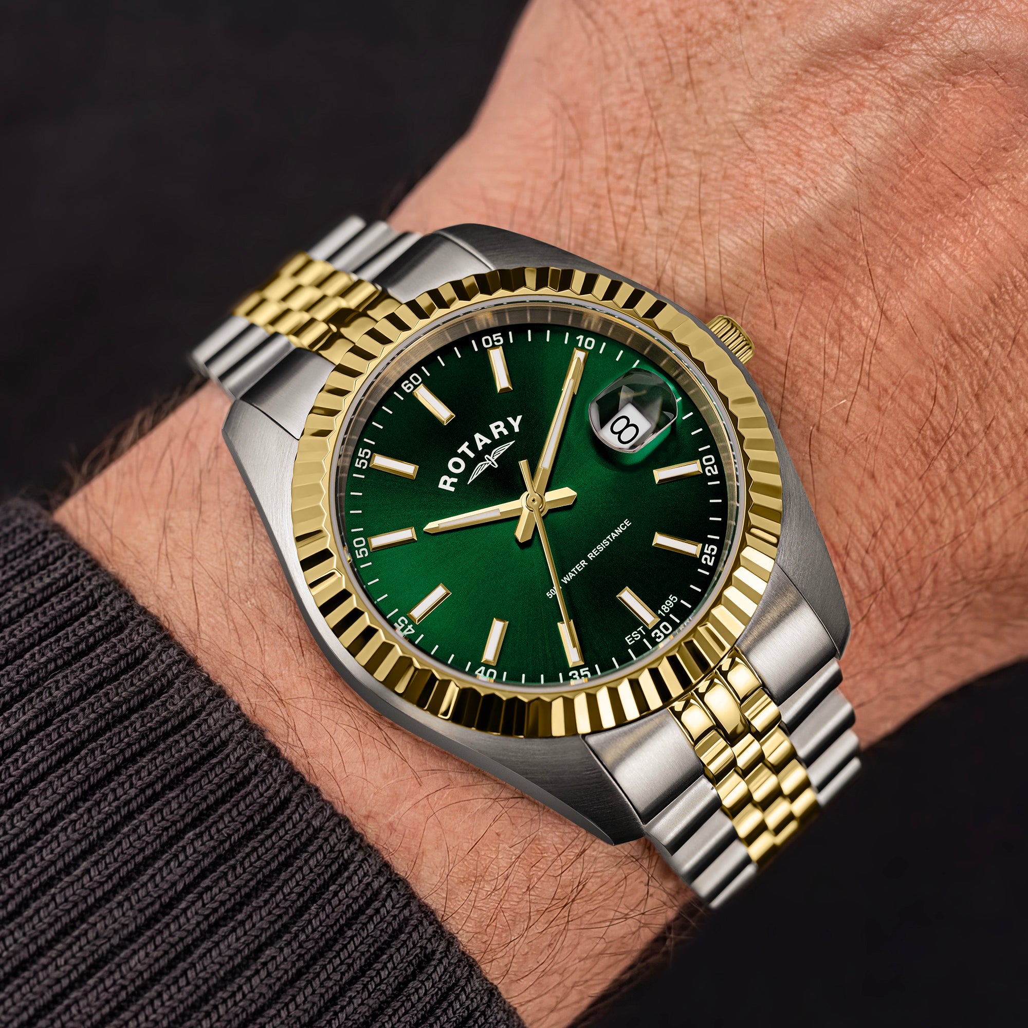 A Rotary wristwatch with a green dial, gold and silver fluted bezel, and a two-tone metal link bracelet, shown worn on a person's wrist.