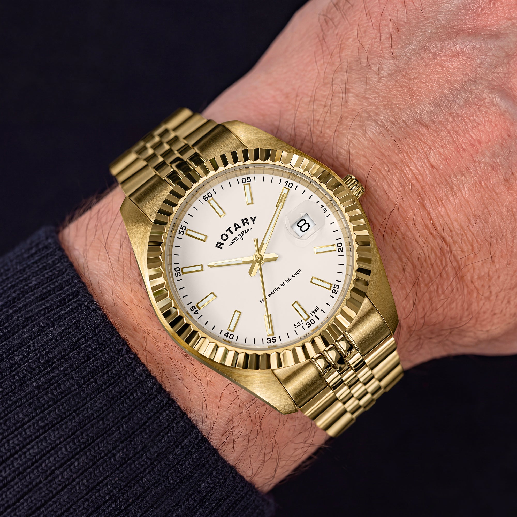 A gold-tone Rotary wristwatch with a white dial and date window, worn on a person's wrist against a dark background.