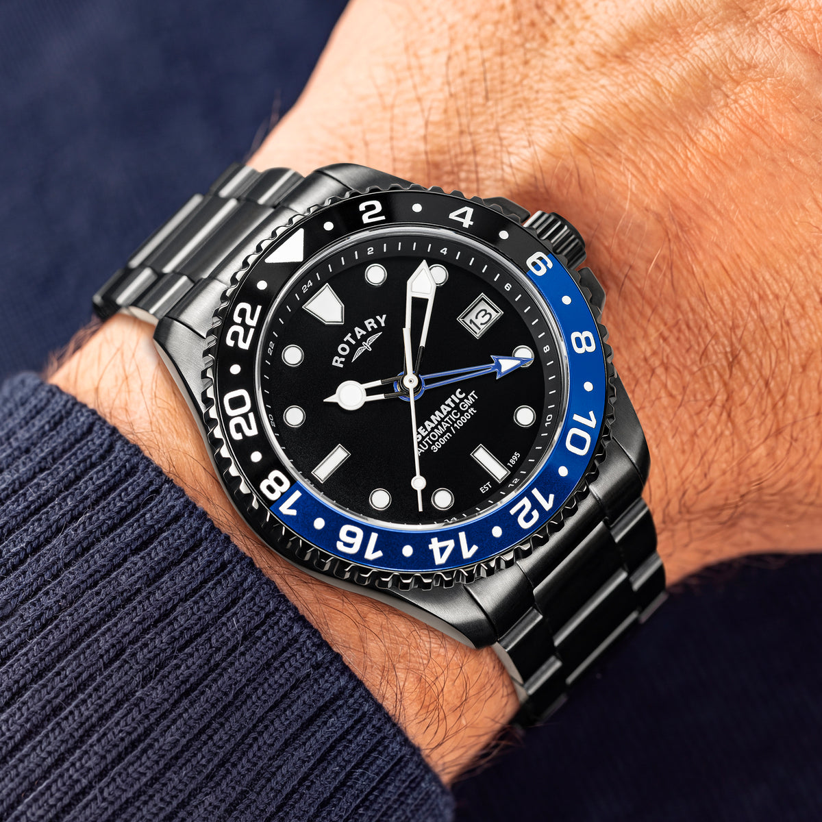 A close-up of a wrist wearing a Rotary Seamatic 300 GMT automatic watch with a black dial, blue bezel section, and black metal bracelet against a dark navy knit sleeve and skin background.