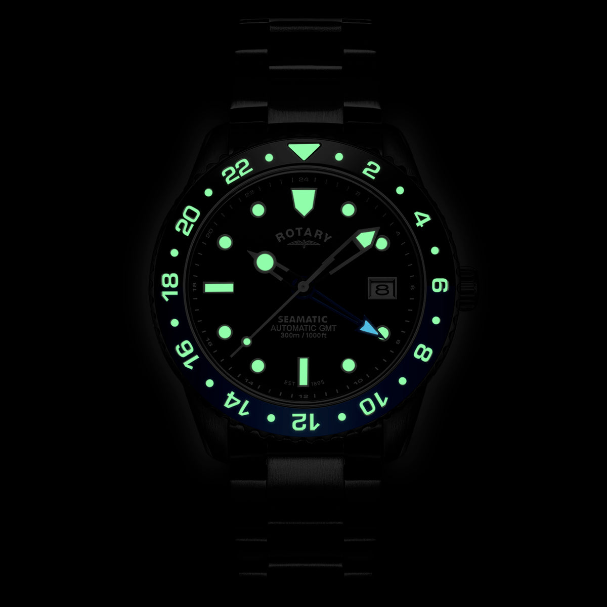 A black Rotary Seamatic 300 GMT wristwatch with a black dial and bezel, illuminated in the dark with glowing green and blue luminescent details. The brand name ROTARY is visible on the dial.