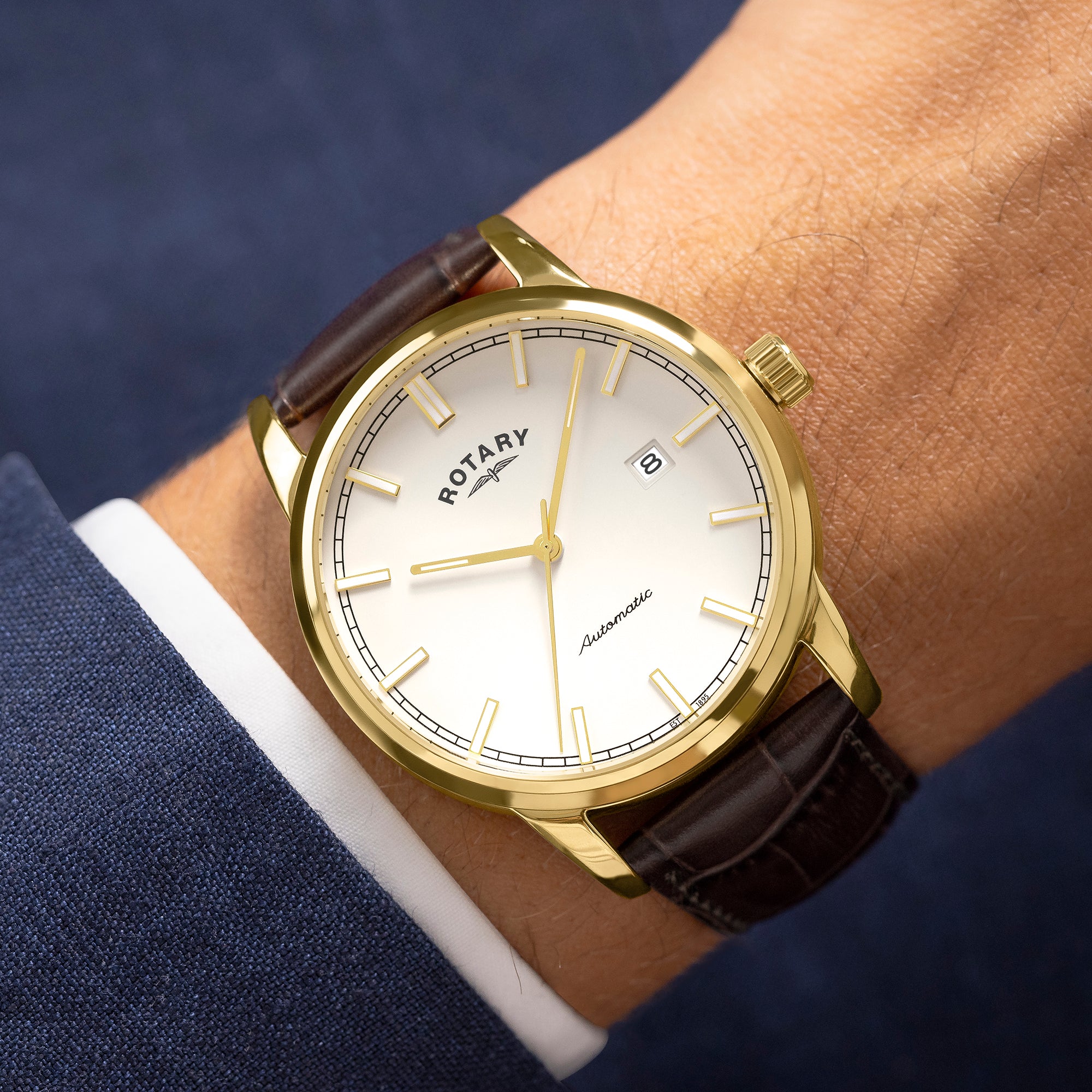 Gold rotary automatic watch with brown leather strap on a wrist against a blue suit background