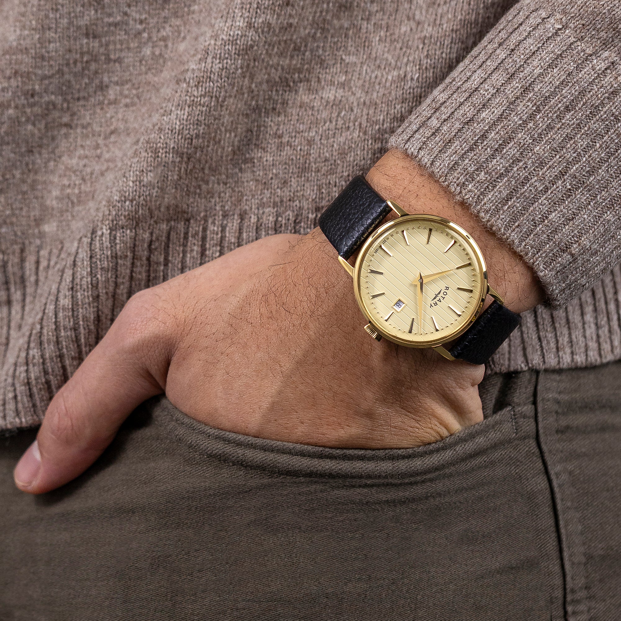Gold Rotary Heritage watch with a black strap on a person's wrist, wearing a brown sweater and gray pants.