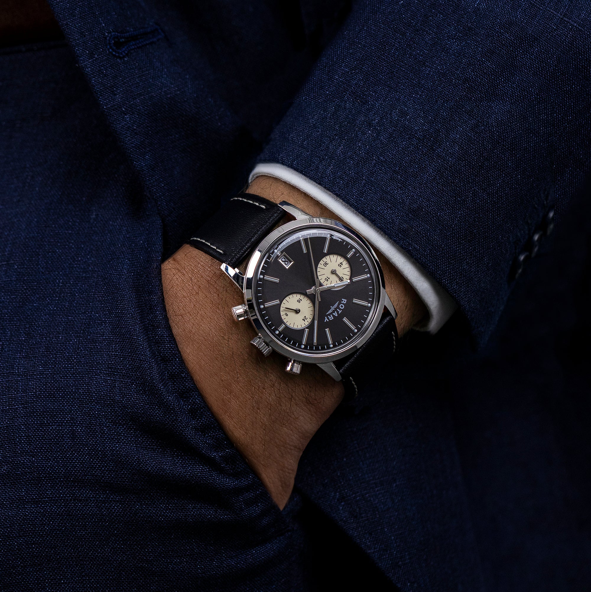 Close-up of a man wearing a dark blue suit with a Rotary 1895 Chronograph wristwatch on a dark background