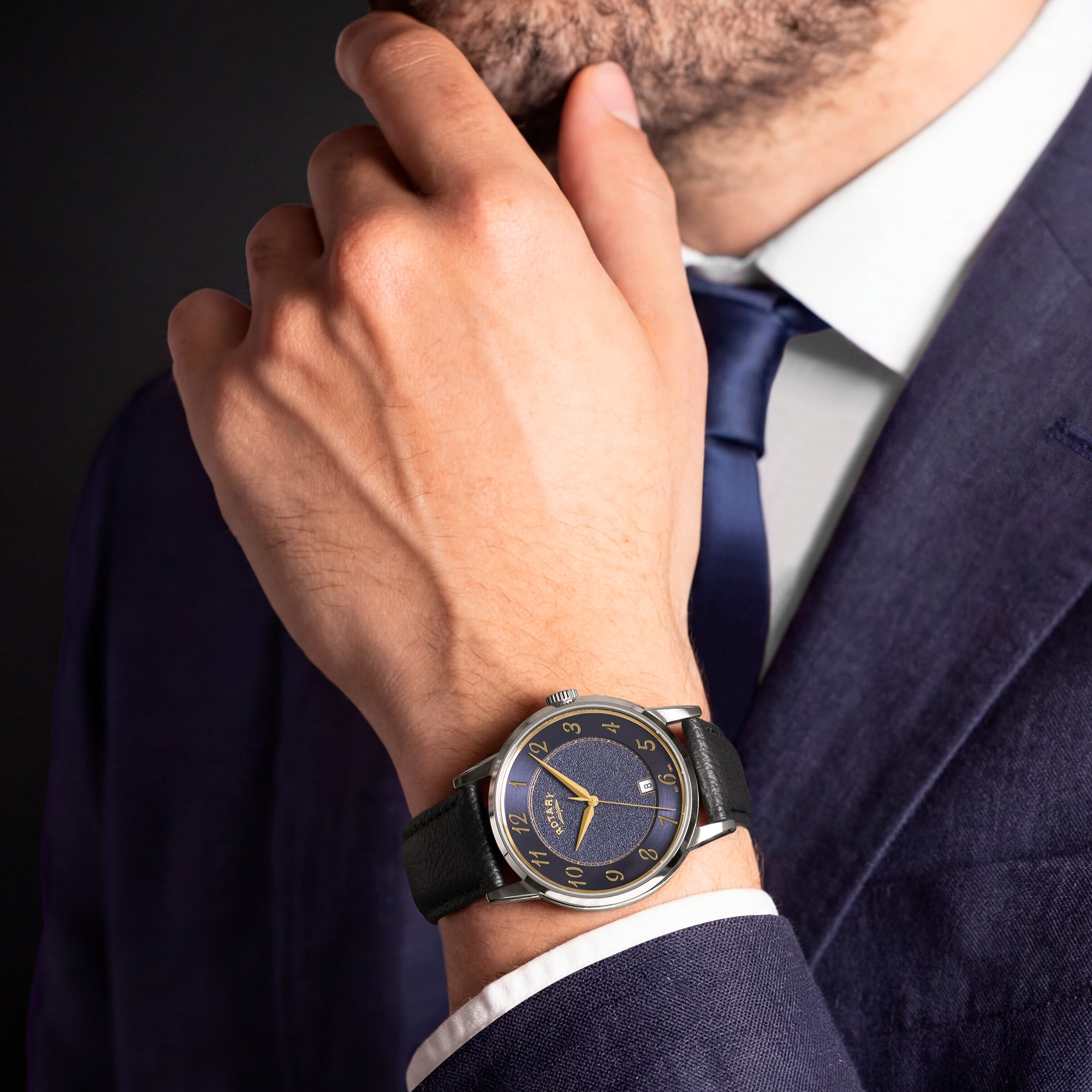 Man wearing a dark suit with a Rotary watch on his wrist against a dark background
