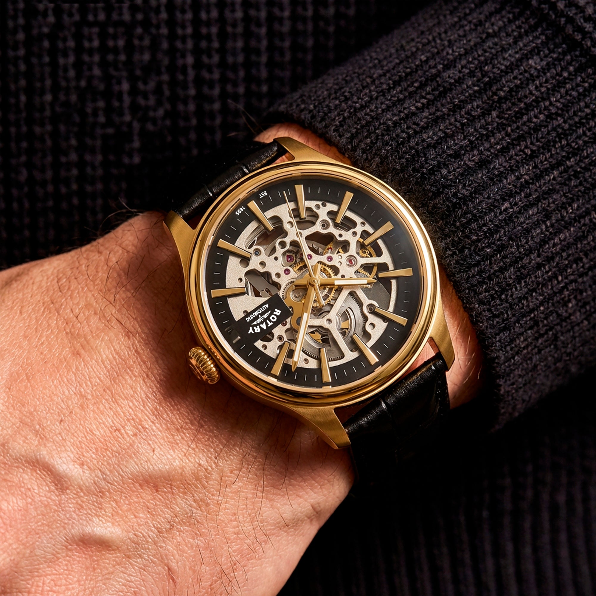 Gold Rotary Skeleton wristwatch with black strap on a person's wrist against a dark background