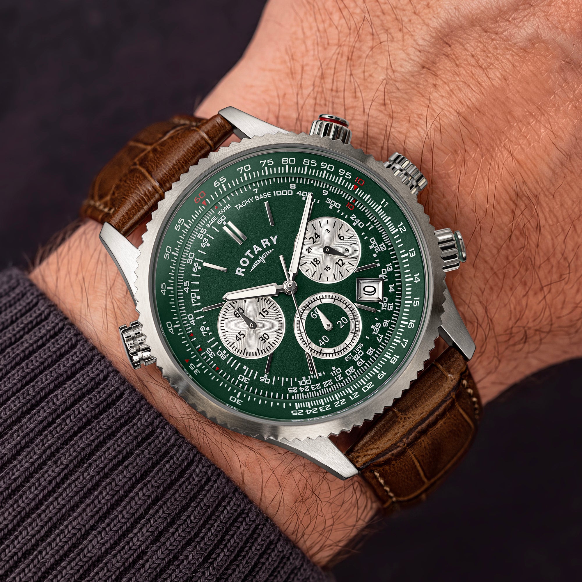 A close-up of a wrist wearing a Rotary Pilot Chronograph watch with a green dial, silver-tone case, and brown leather strap against a dark background.