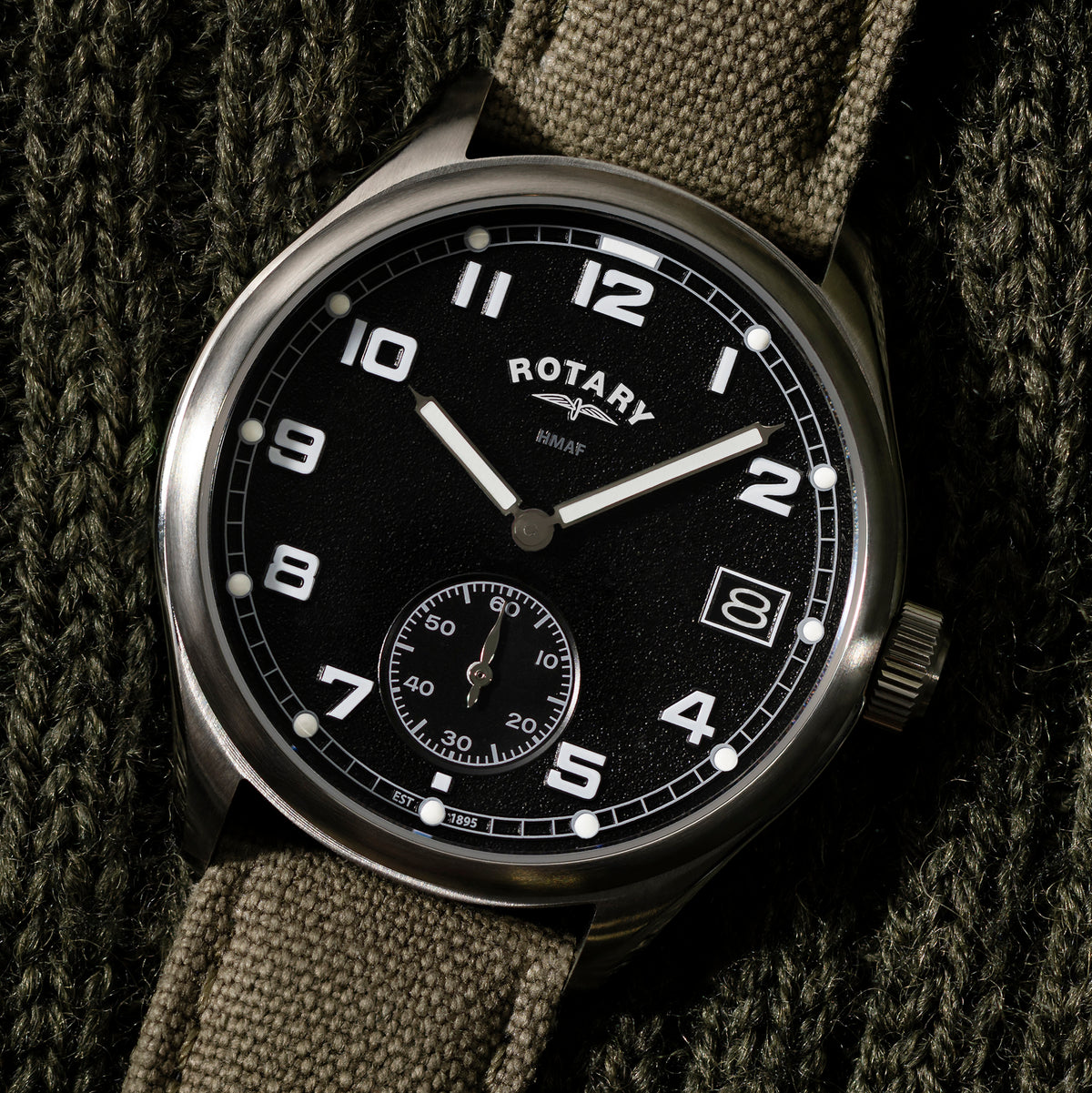 A close-up of a Rotary x MoD British Army wristwatch with a black dial, silver-tone case, and olive green canvas strap, resting on a dark textured knit background.