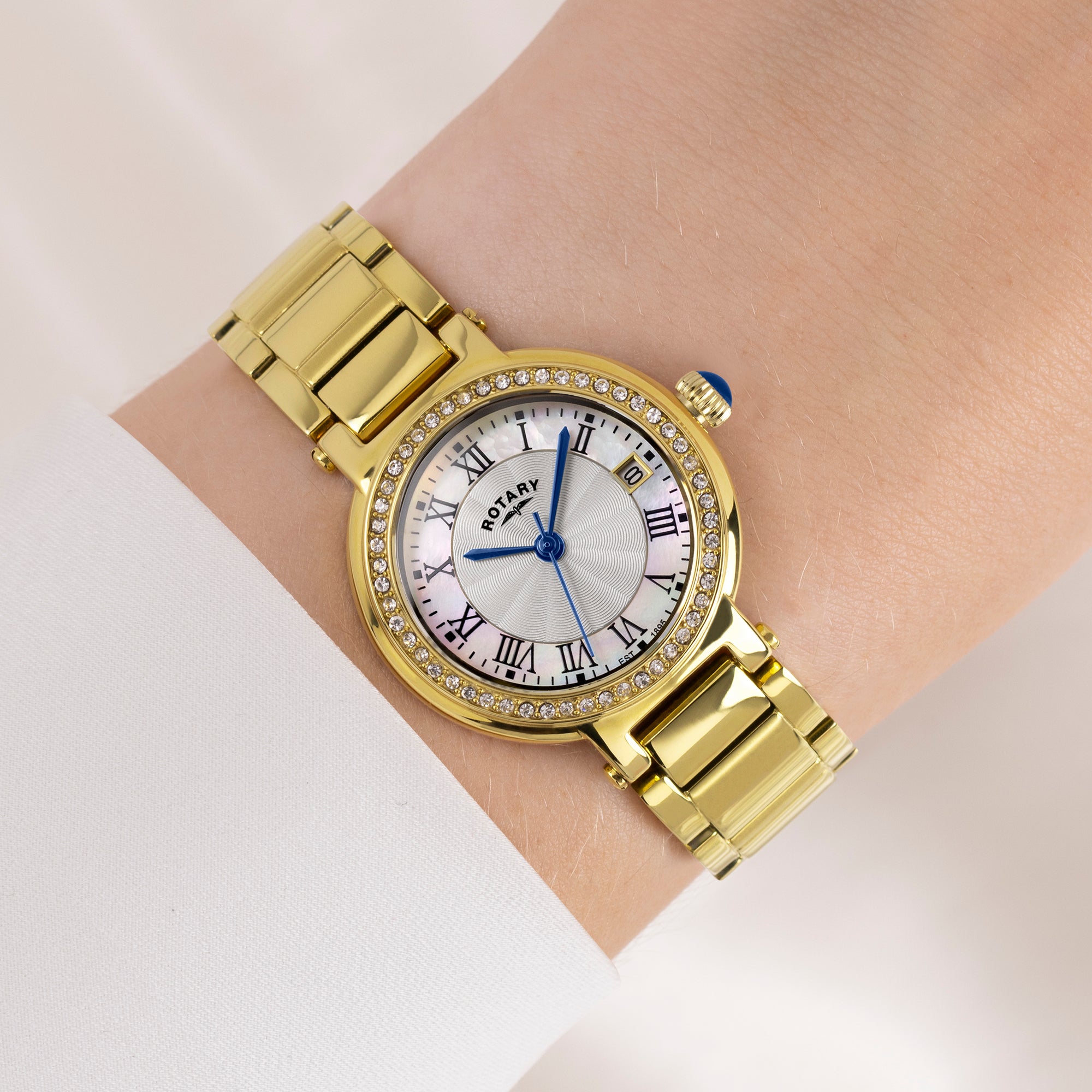 Gold wristwatch with a white face and blue hands on a wrist against a light background
