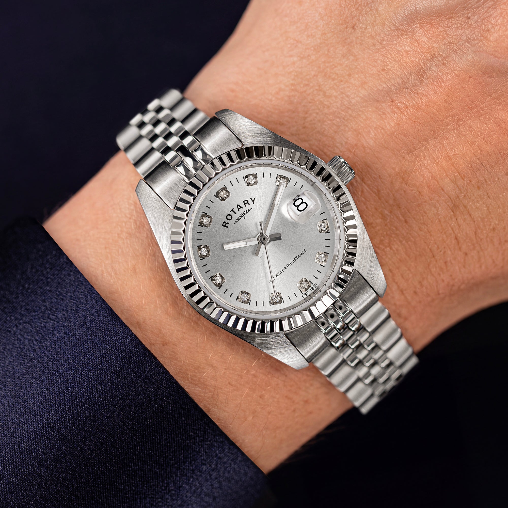 A silver Rotary stainless steel wristwatch with a fluted bezel, silver dial, and matching metal link bracelet, shown worn on a person's wrist against a dark navy sleeve.
