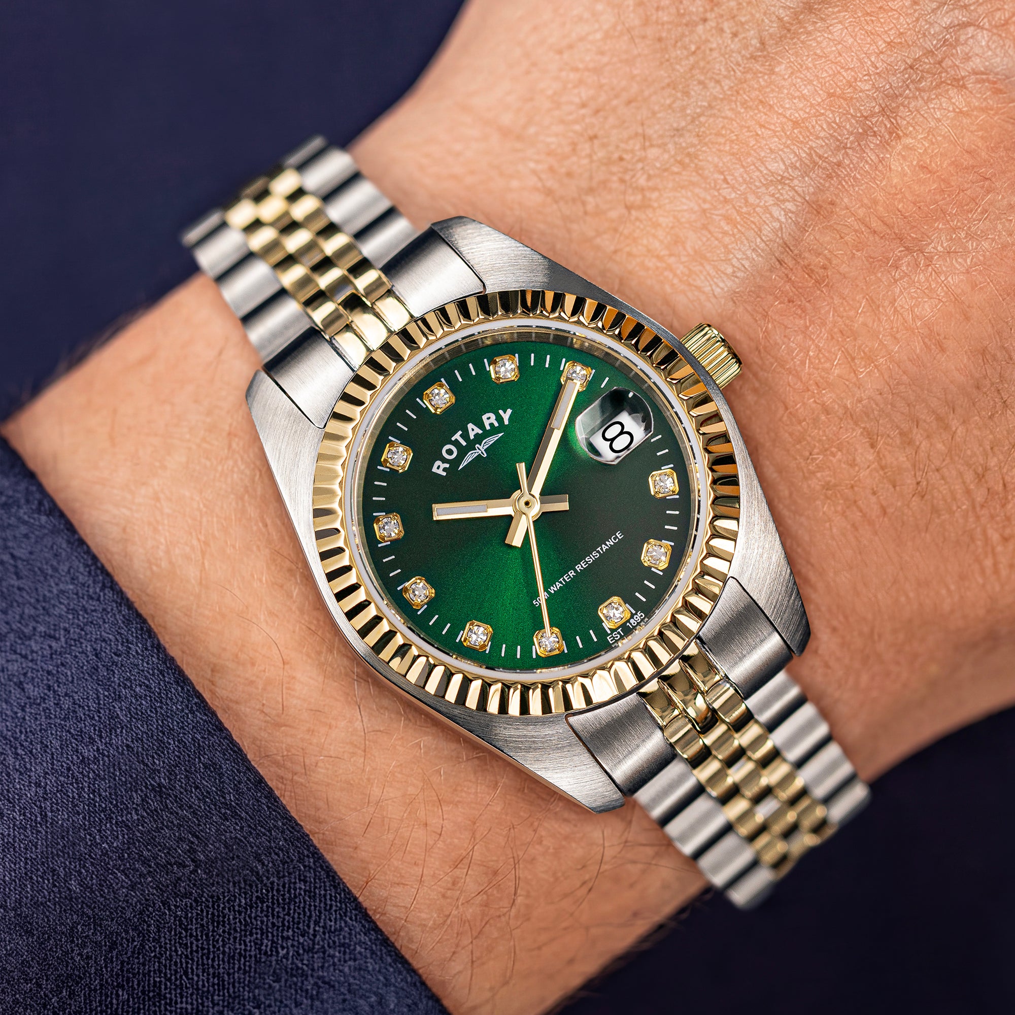 A close-up of a wrist wearing a two-tone Rotary watch with a green dial and gold-tone accents. The watch has a fluted bezel, gold-tone hour markers, and a date window at the 3 o'clock position.
