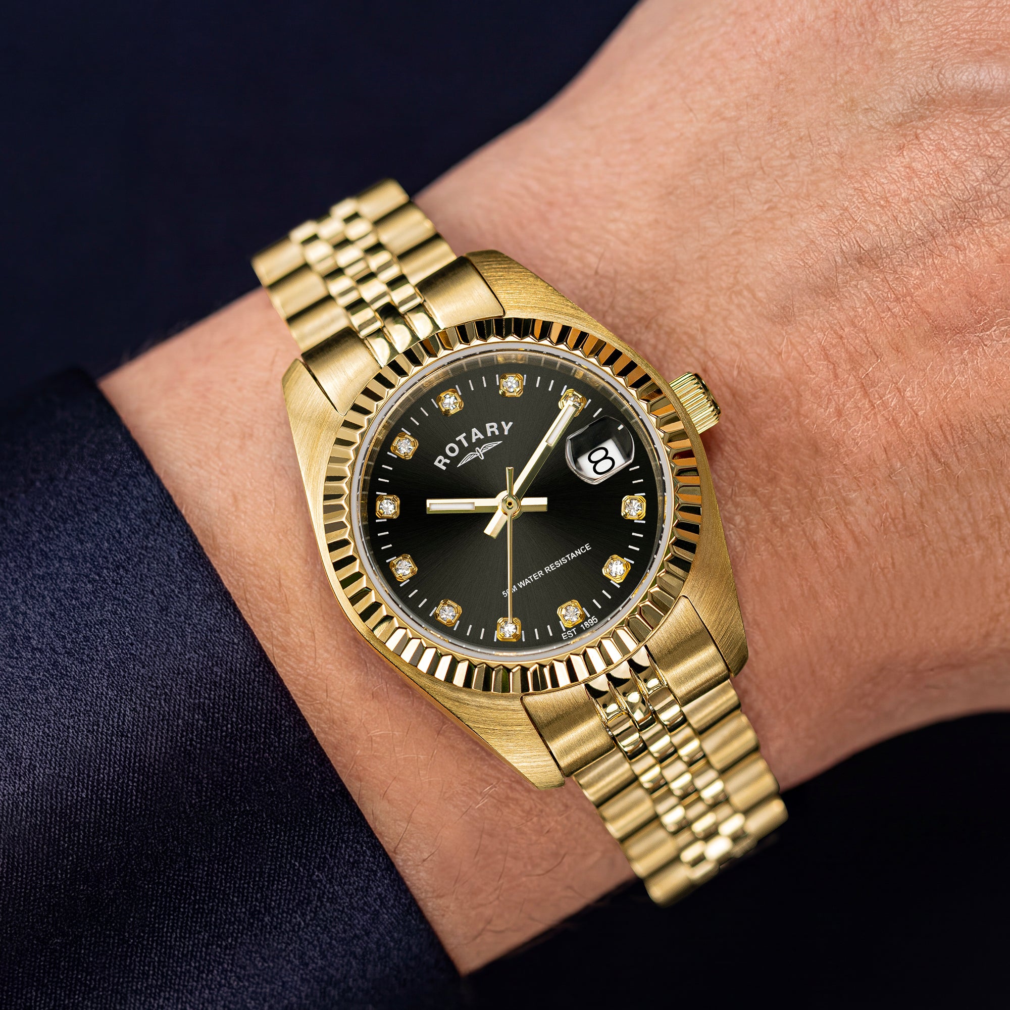 A gold-tone Rotary ladies wristwatch with a black dial and diamond hour markers, shown worn on a person's wrist against a dark navy sleeve background.