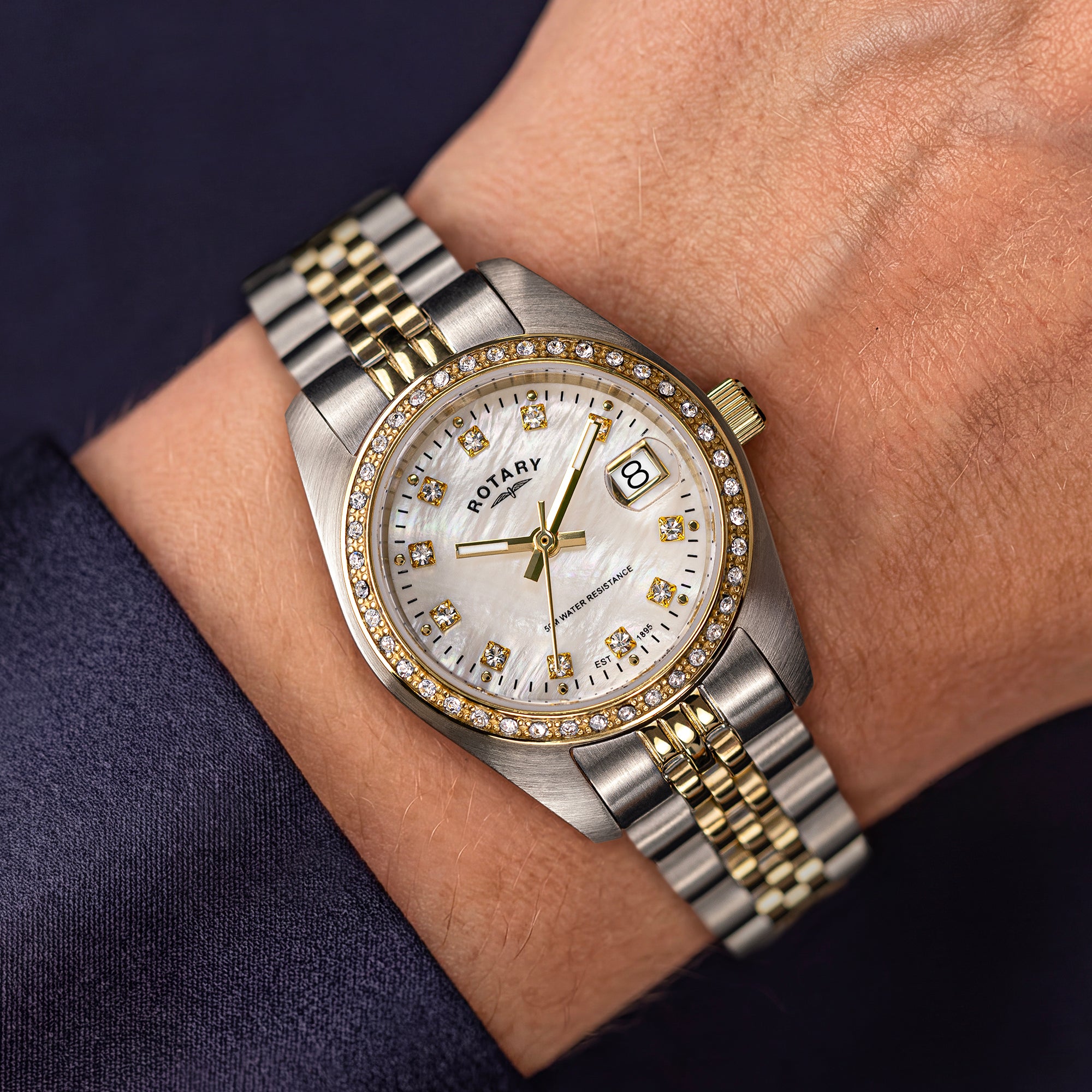 A close-up of a wrist wearing a Rotary Dress Date watch with a two-tone stainless steel and gold bracelet, a white dial, and a bezel set with diamonds, against a dark navy sleeve and skin background.