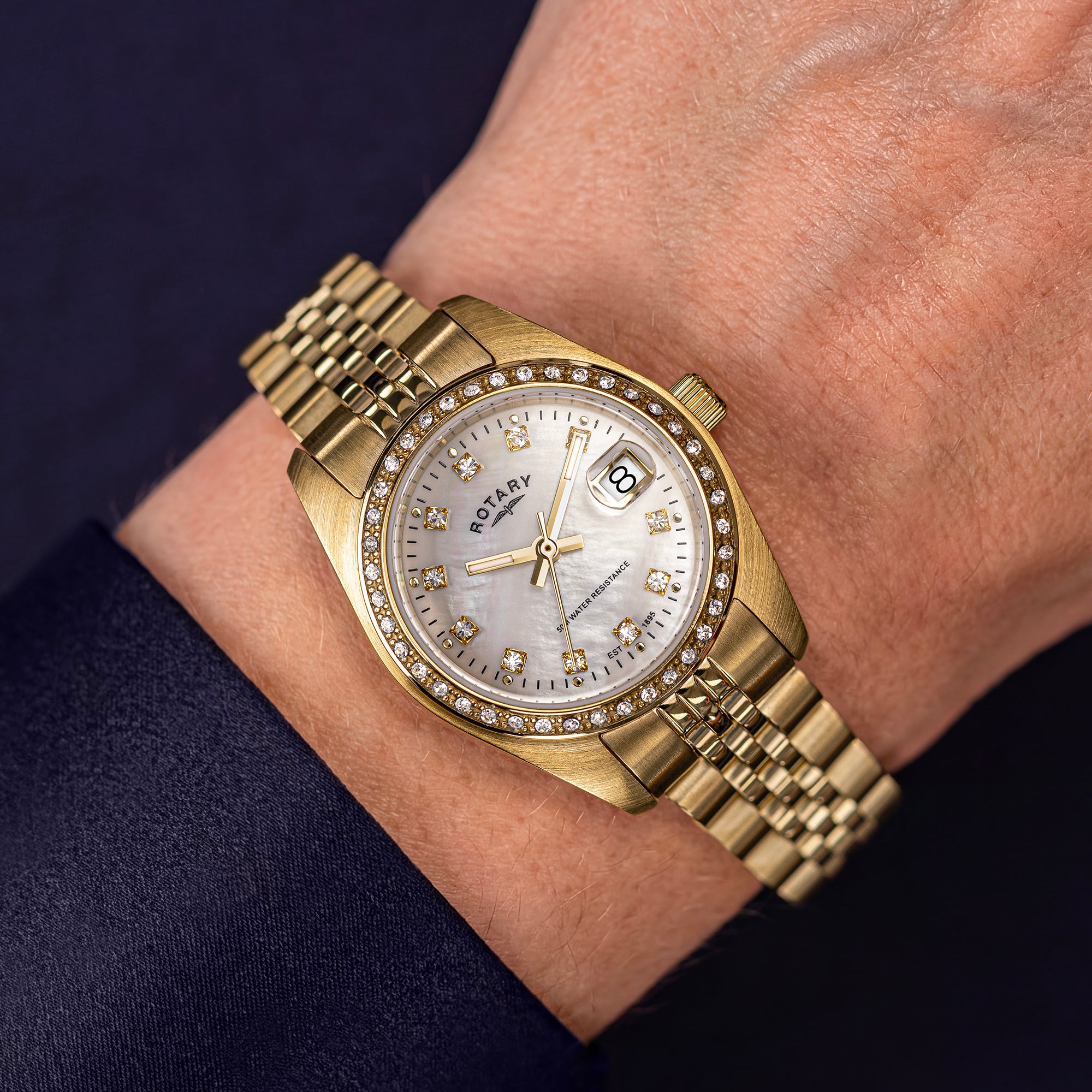 A gold-tone Rotary Dress Date watch with a diamond-accented bezel and a silver-tone mother-of-pearl dial, shown worn on a wrist against a dark navy background.