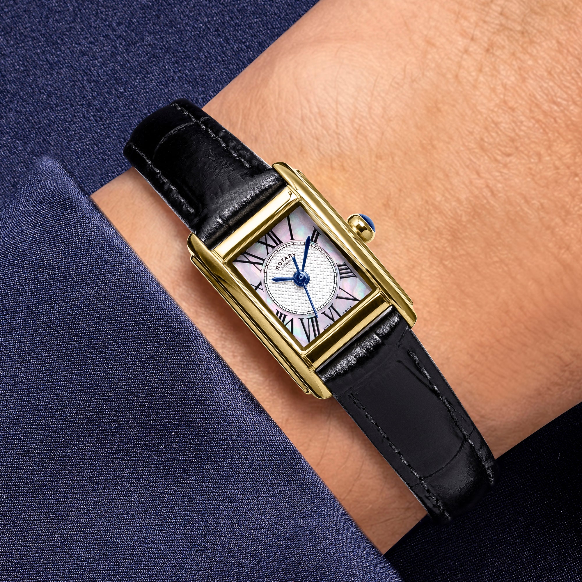 Gold Rotary Heritage Astoria watch with black leather strap on a wrist against a dark blue fabric background