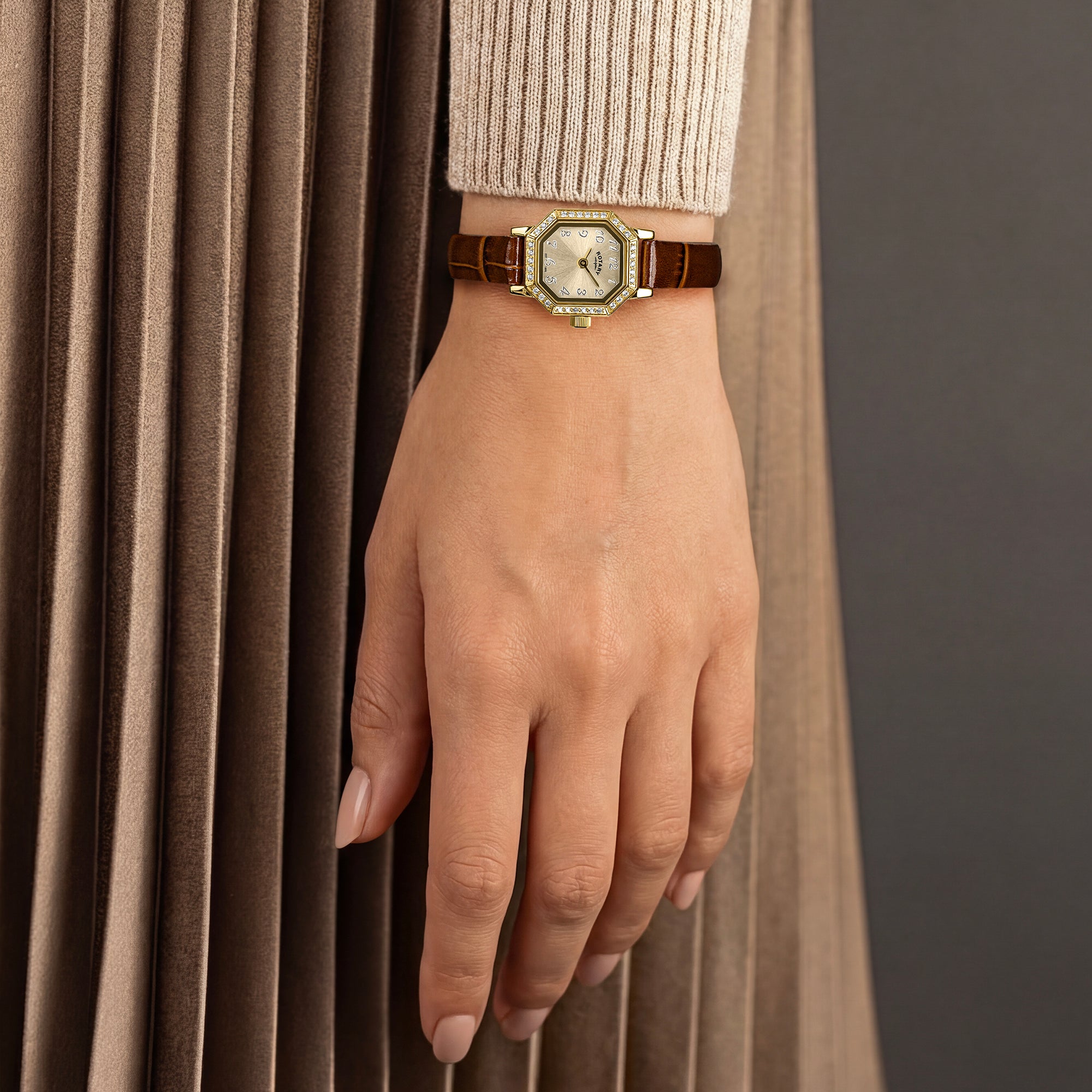 Women's hand wearing a vintage inspired gold Rotary watch with a brown strap against a neutral background