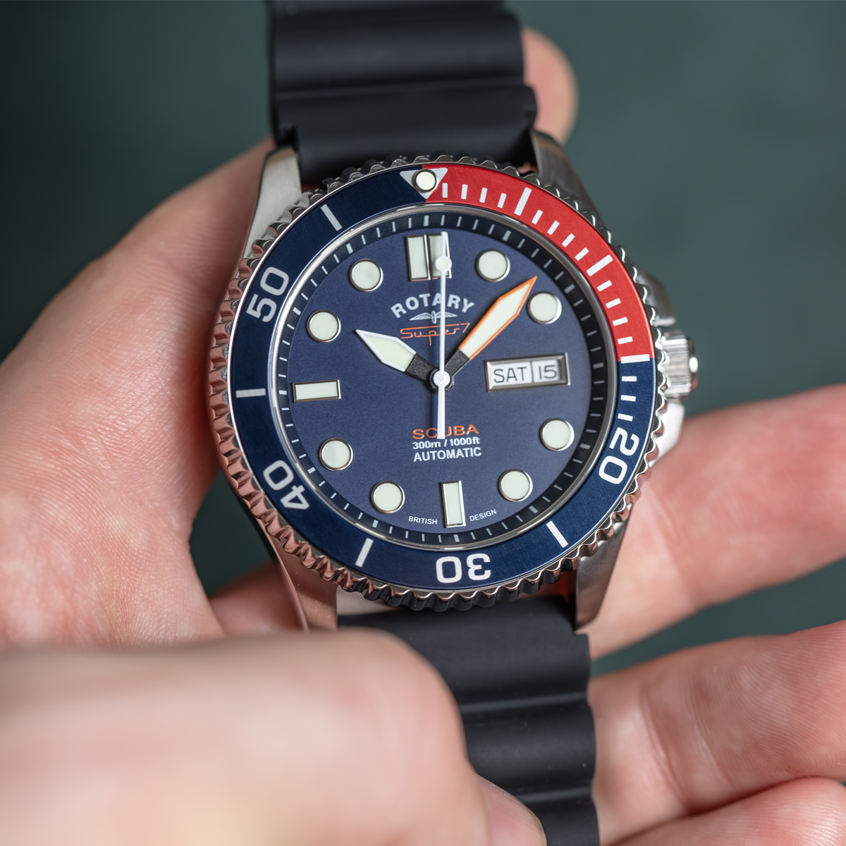 Close-up of a Rotary watch with a blue and red bezel held in a hand against a dark background.