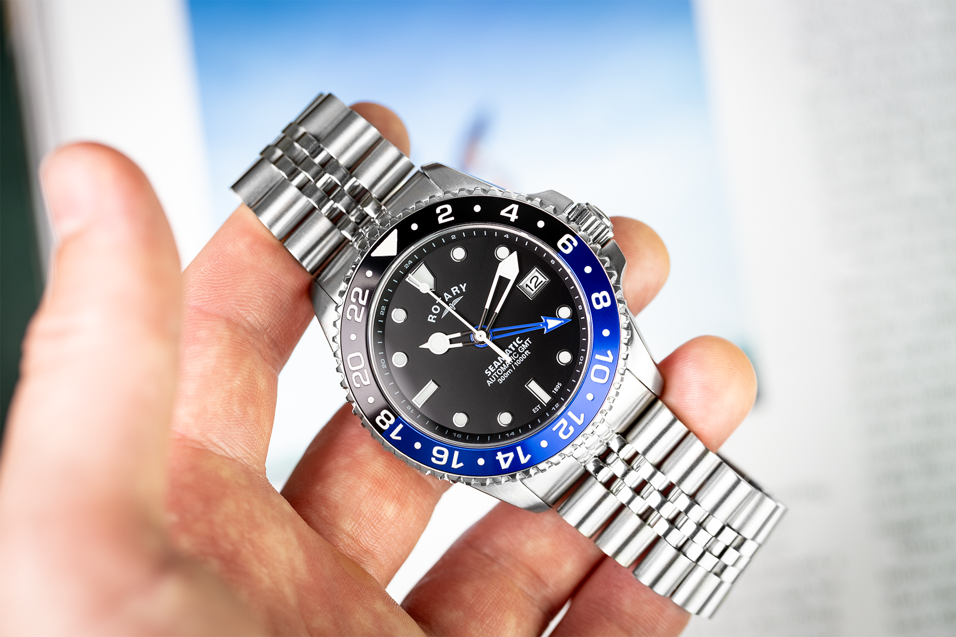 A Rotary Seamatic 300 GMT wristwatch with a silver metal bracelet and a black dial, held in a hand. The bezel is black and blue with white inserts.
