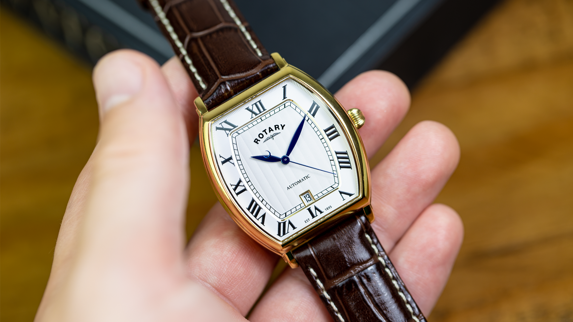 A close-up of a hand holding a gold-tone Rotary 1895 Tonneau watch with a brown leather strap and a white dial featuring Roman numerals. The brand name 'ROTARY' is visible on the watch face.