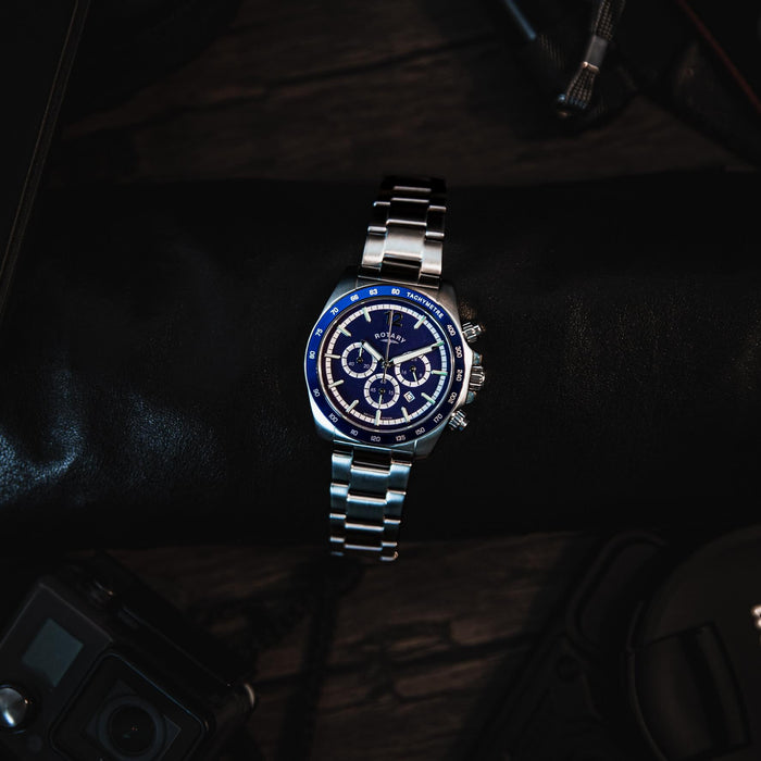 A stainless steel rotary chronograph watch with a blue dial and bezel, lying on a table in a moody setting