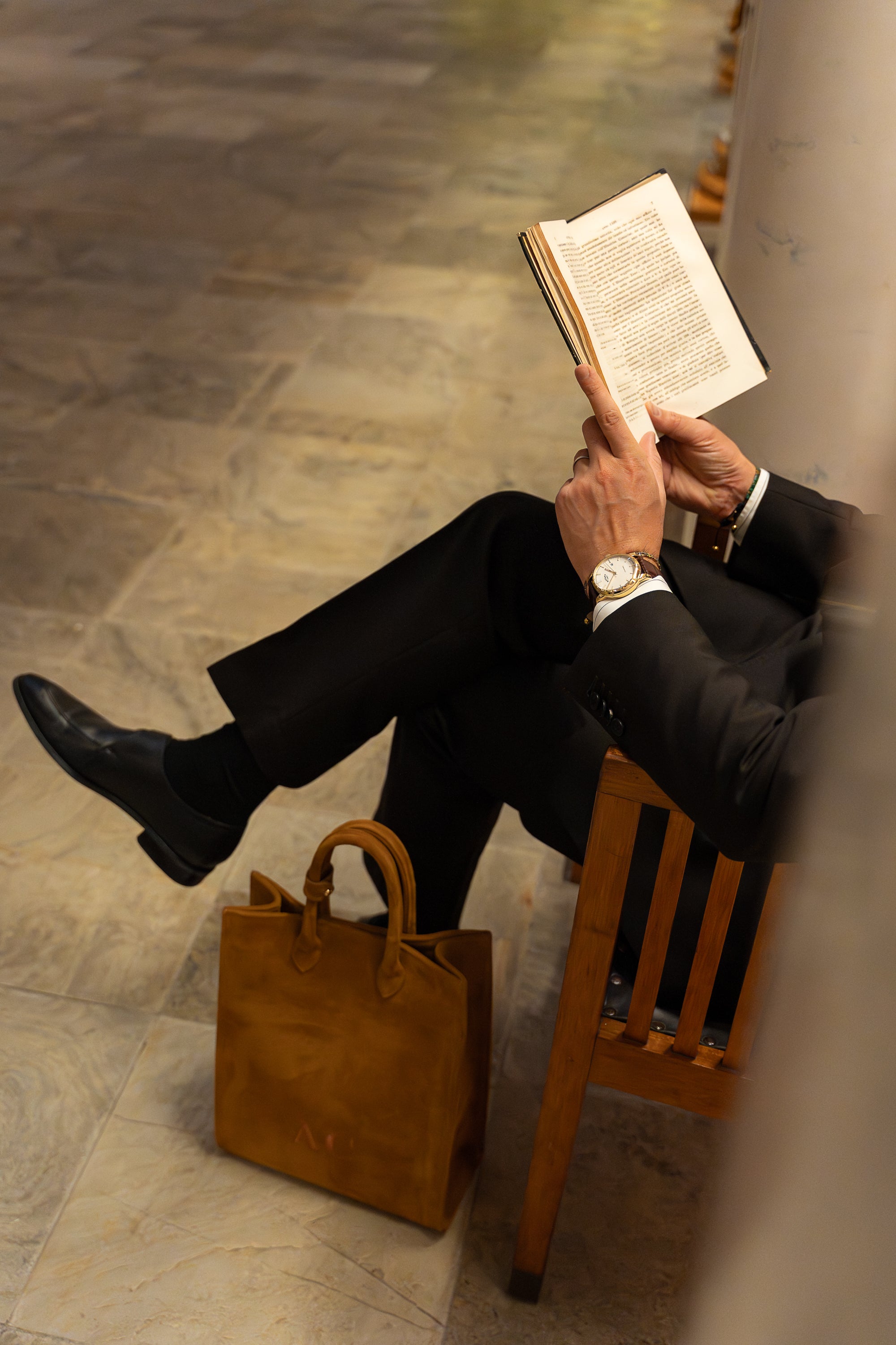 Man in a suit wearing a gold rotary watch reading a book with a brown bag beside him on a stone floor.