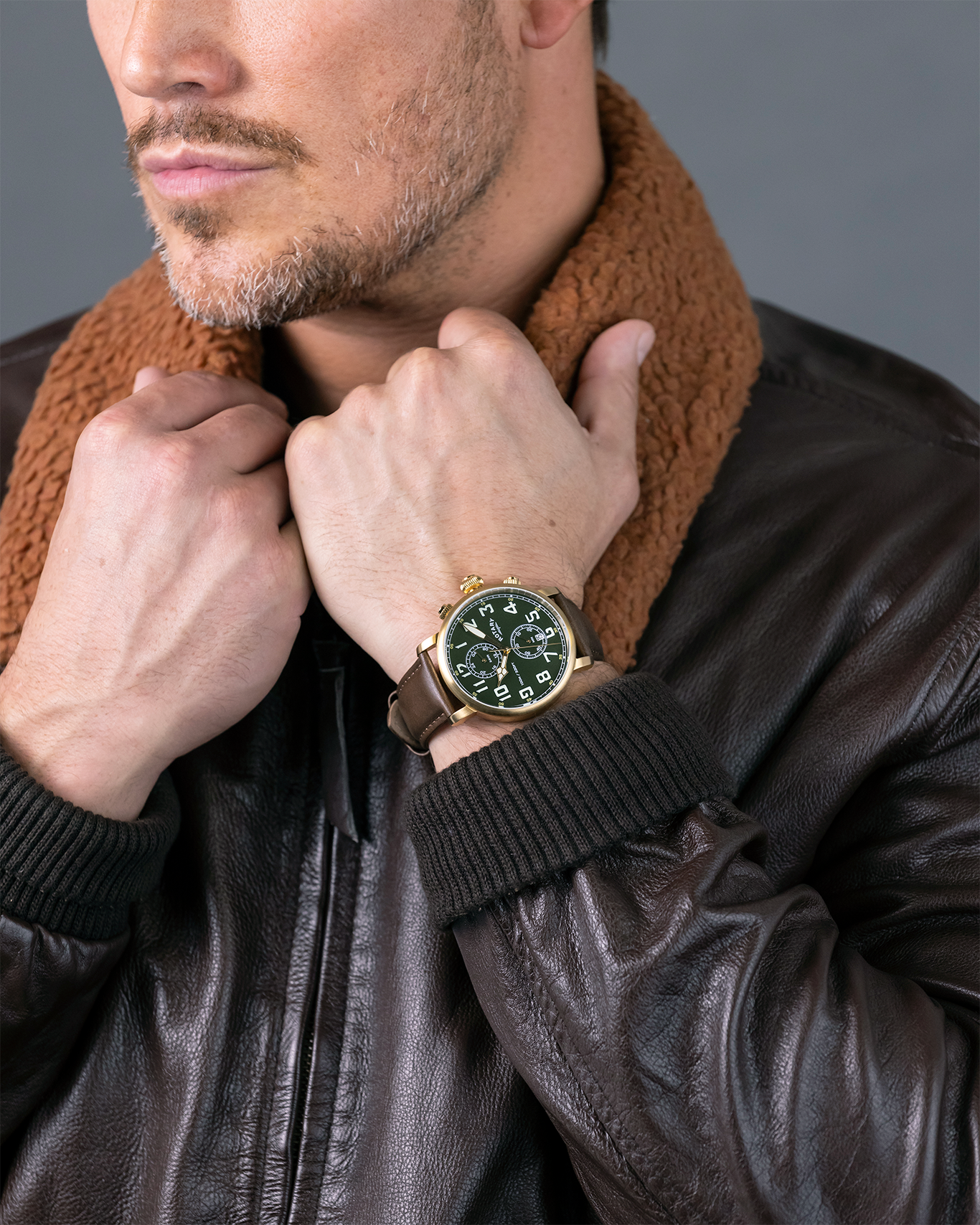 Man wearing a brown leather jacket and brown scarf with a Rotary Apex100 Pilot watch on his wrist against a gray background