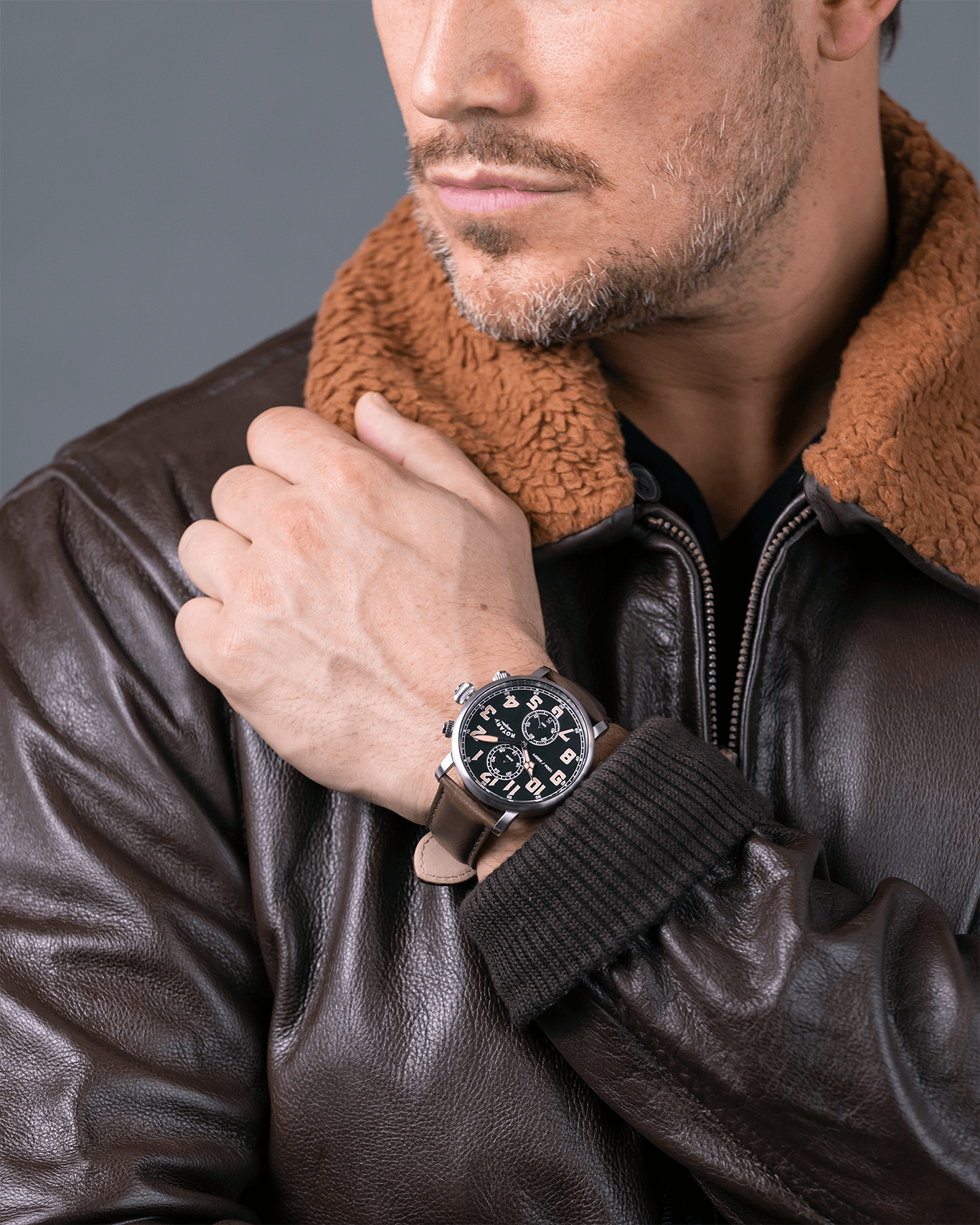 Man wearing a brown leather jacket with a fur-lined hood, close-up of his wrist with a Rotary Apex100 Pilot watch.