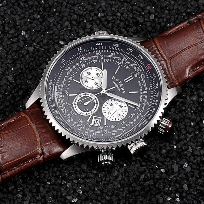 a close up of a Rotary pilot style watch with a stainless steel case and a brown leather strap. The watch has a date window and 3 chronograph sub-dials sitting on a gravel background