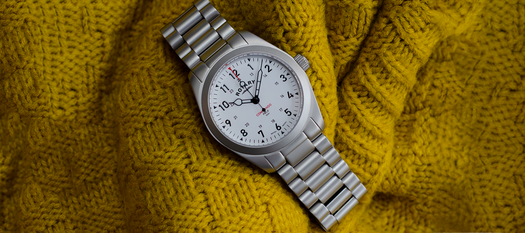 rotary mens watch in stainless steel with a white dial and three hands. The watch is sitting on a yellow textured background