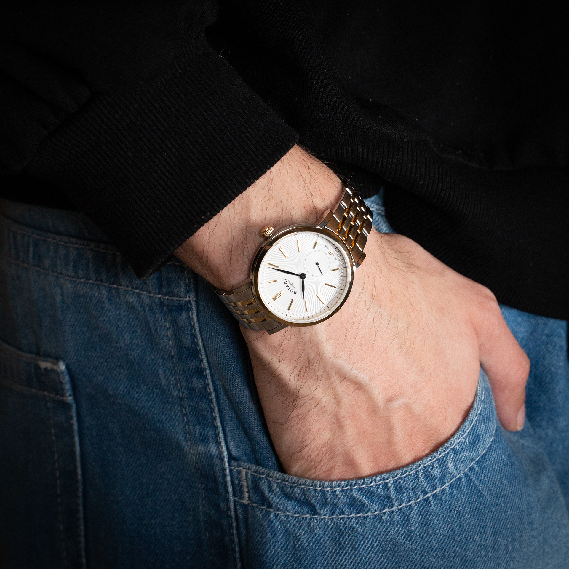 Rotary mens watch in gold and stainless steel with a white dial and blue hands. The watch is being worn by a man wearing jeans and a black jumper