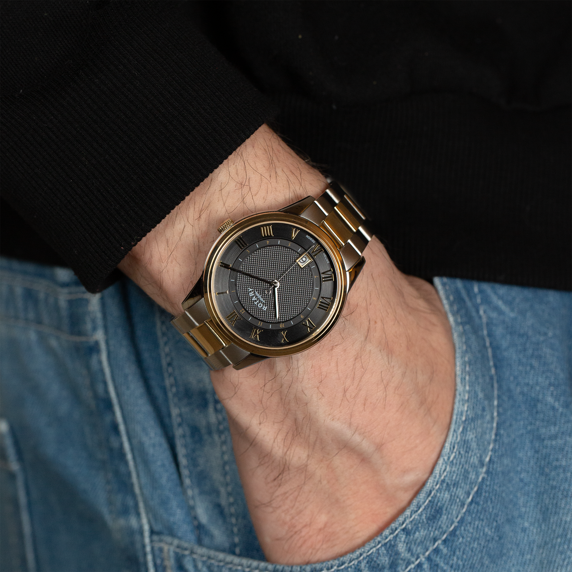 A man with his hand in his pocket, wearing a Rotary mens watch in two tone stainless steel and gold. The watch has a grey dial with traditional style roman numerals in gold