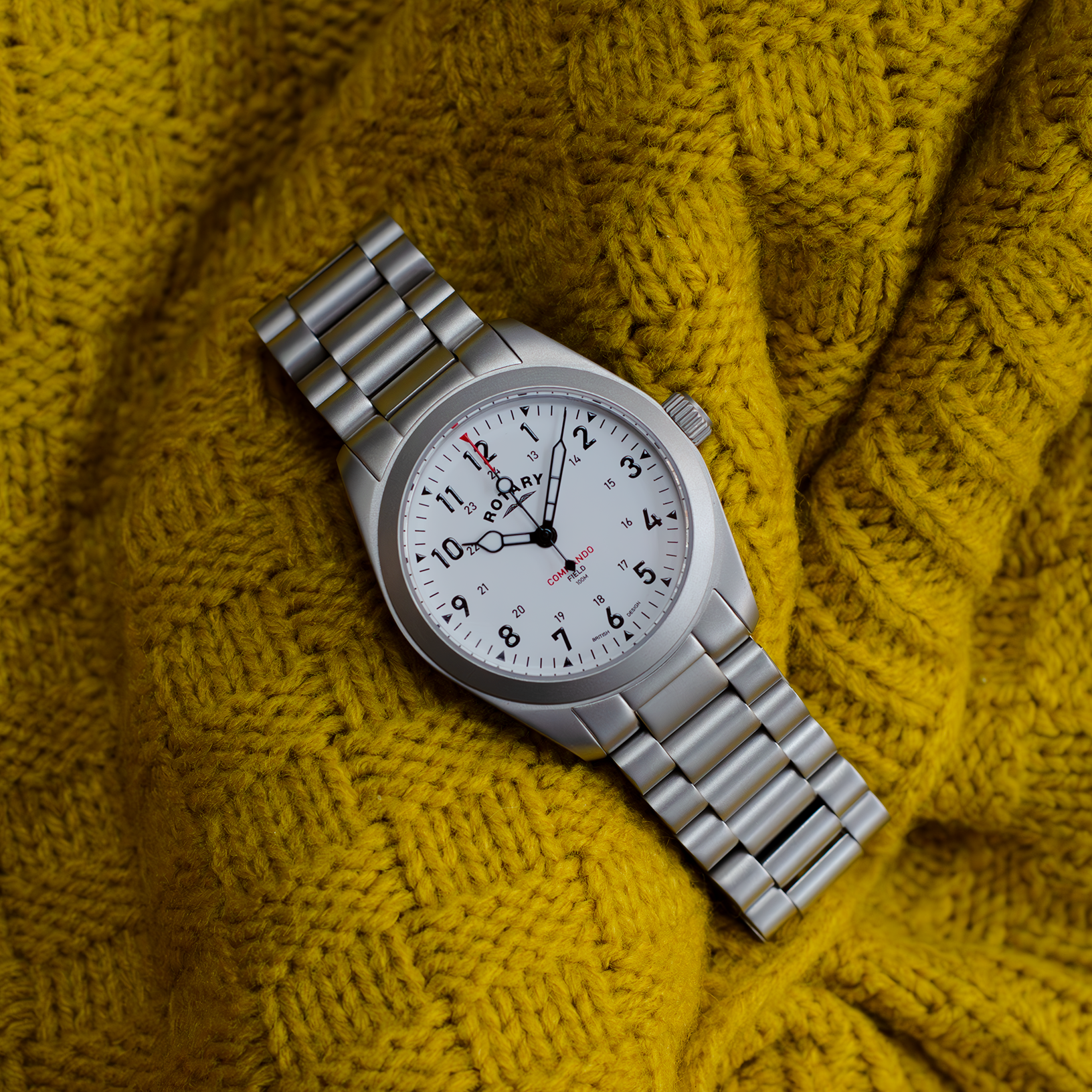 rotary mens watch in stainless steel with a white dial and three hands. The watch is sitting on a yellow textured background