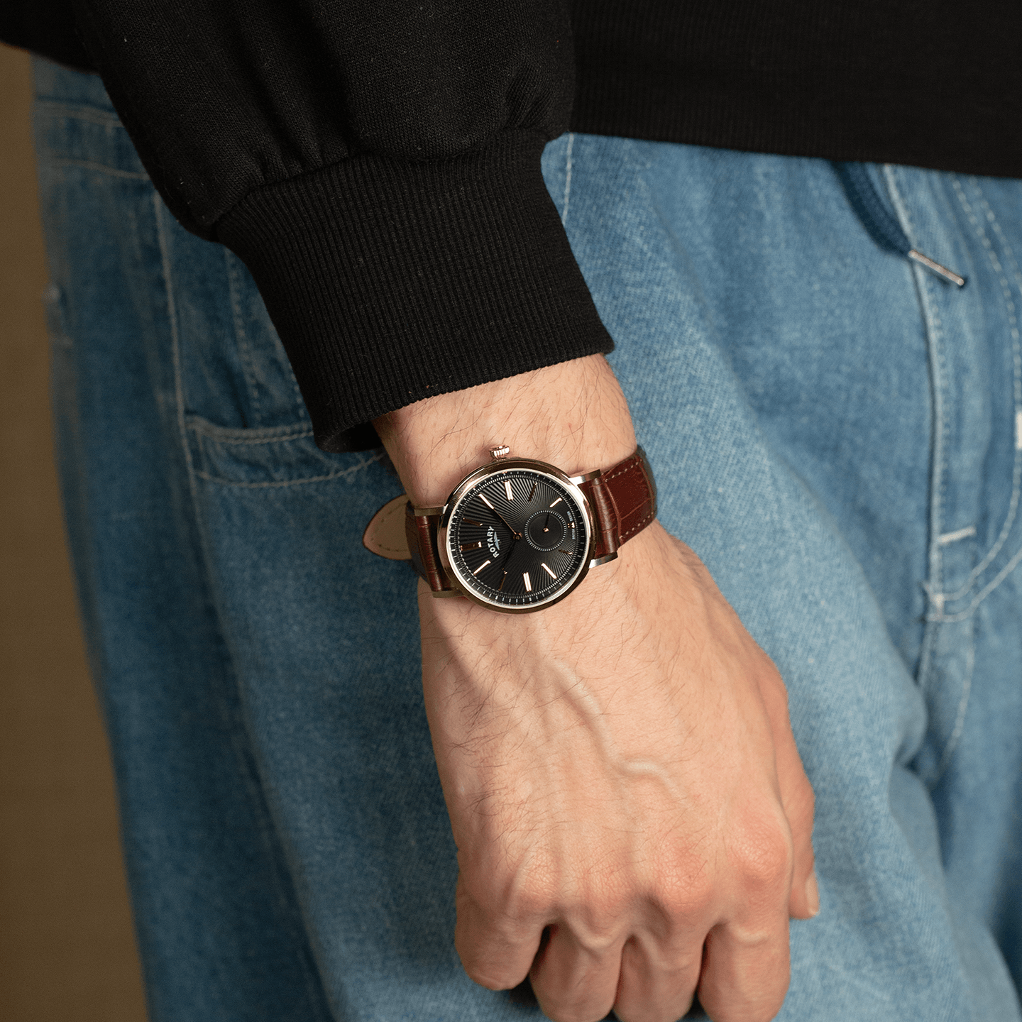 stainless steel rotary watch with a brown leather strap being worn by a man with a black top and jeans