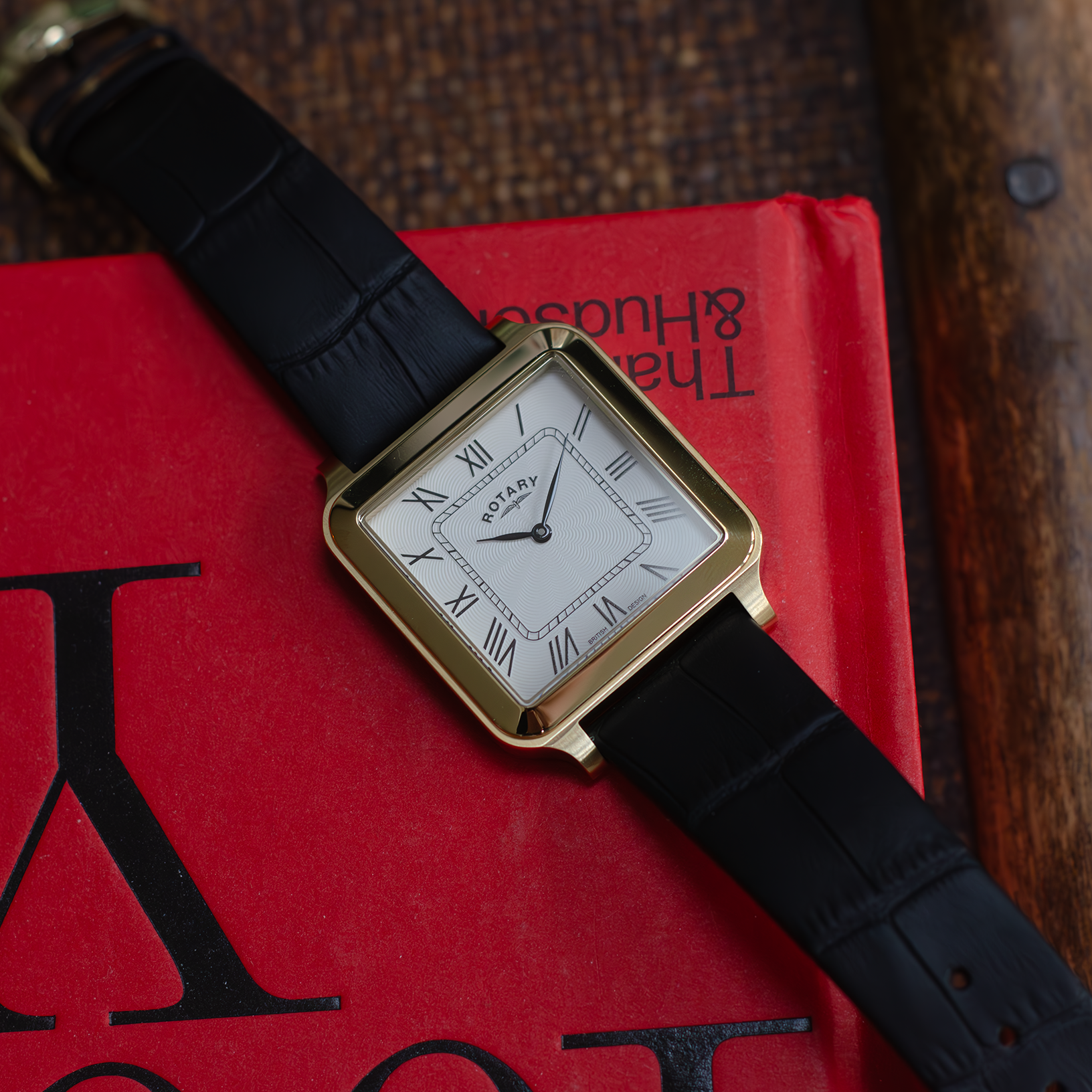 A mens rotary watch in gold with a black leather strap lying on a red book with a brown wooden background