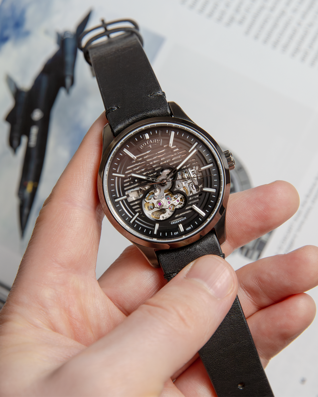 Hand holding a black Rotary Greenwich GT1 wristwatch with a detailed face against a blurred background