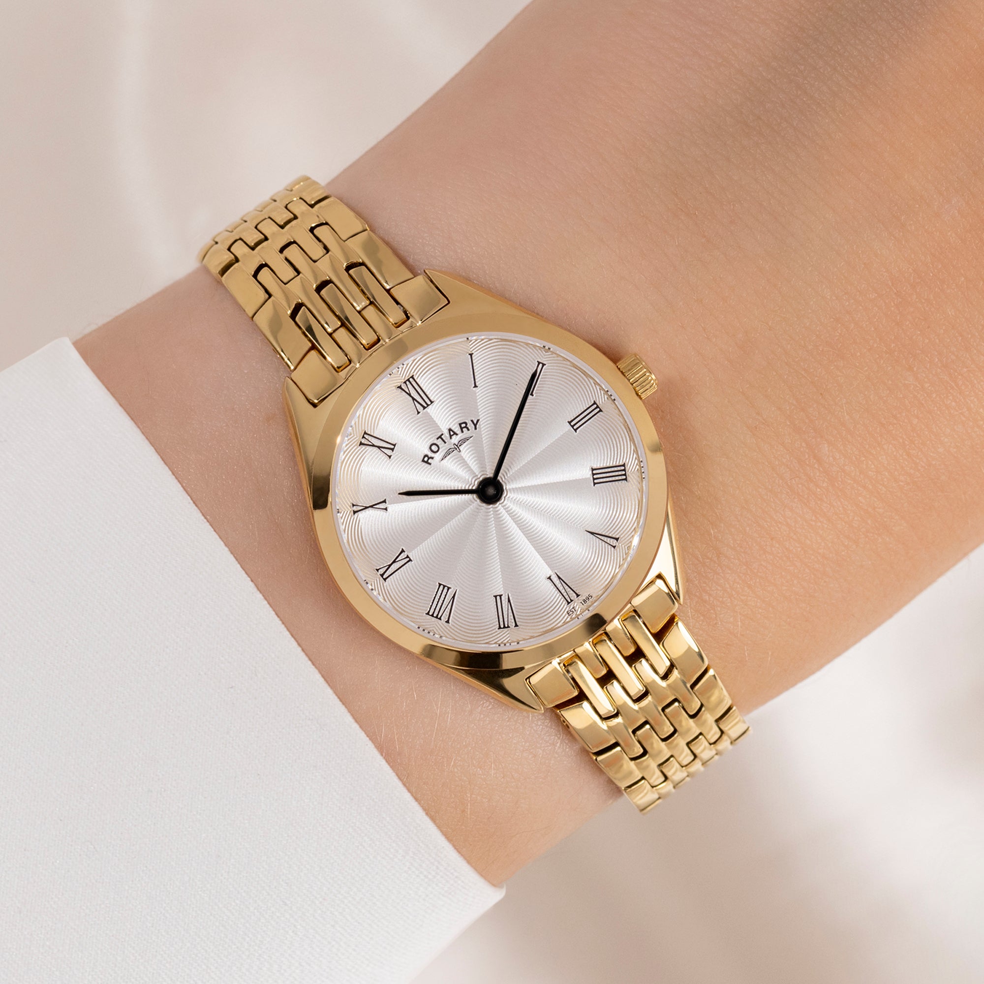 Gold Rotary wristwatch with a white face and Roman numerals on a wrist against a light background