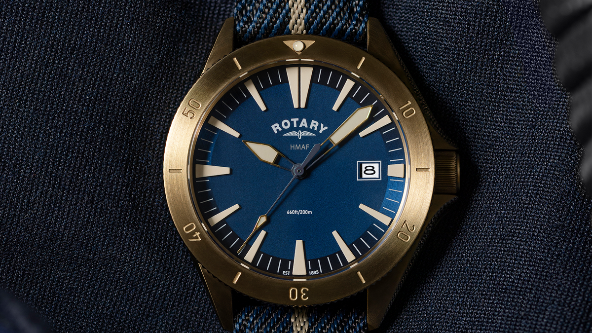 A close-up of a Rotary x MoD Royal Navy watch with a blue dial, gold-tone case, and blue-and-white striped fabric strap, resting on dark denim.