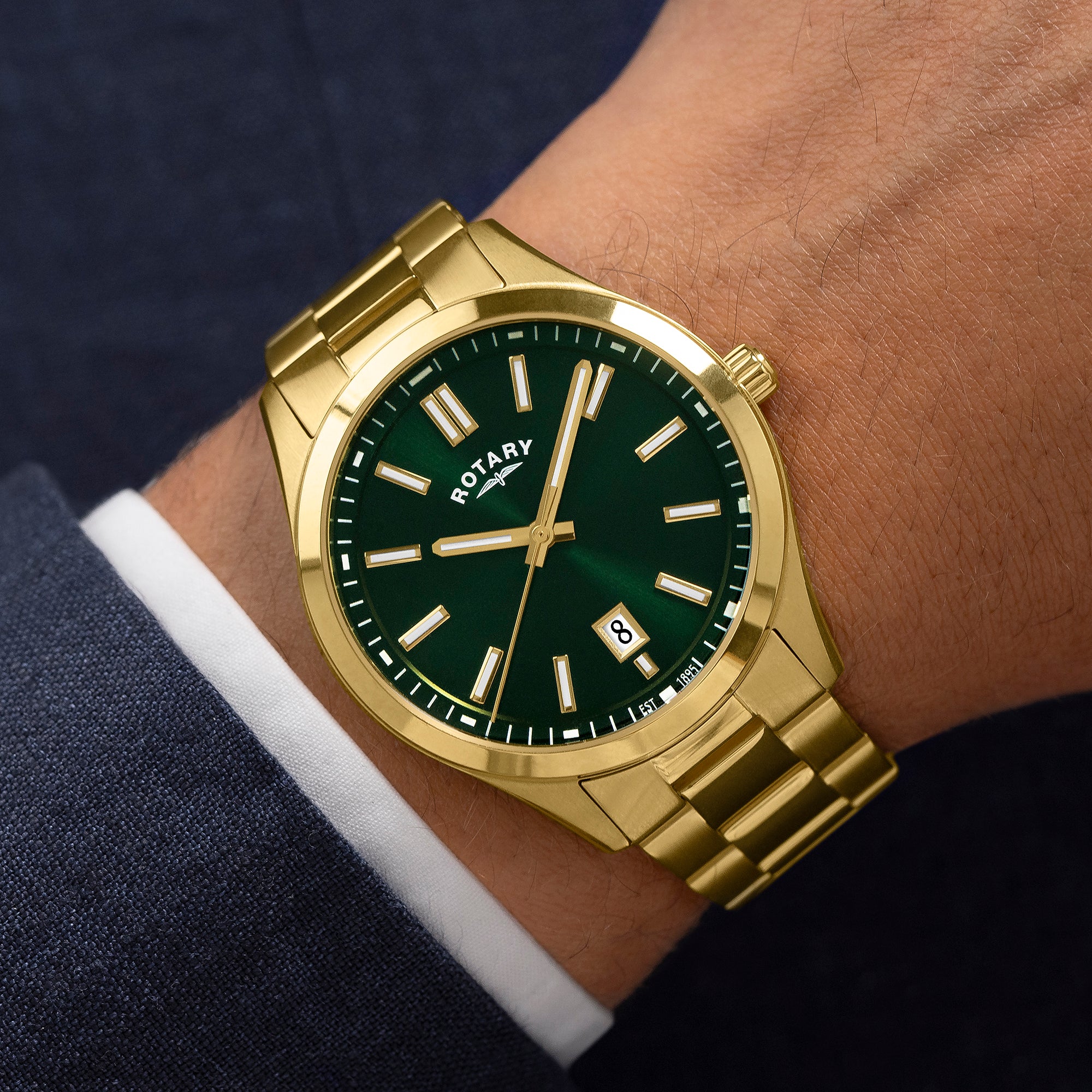 Gold Rotary wristwatch with a green dial on a person's wrist, wearing a dark suit and white shirt.