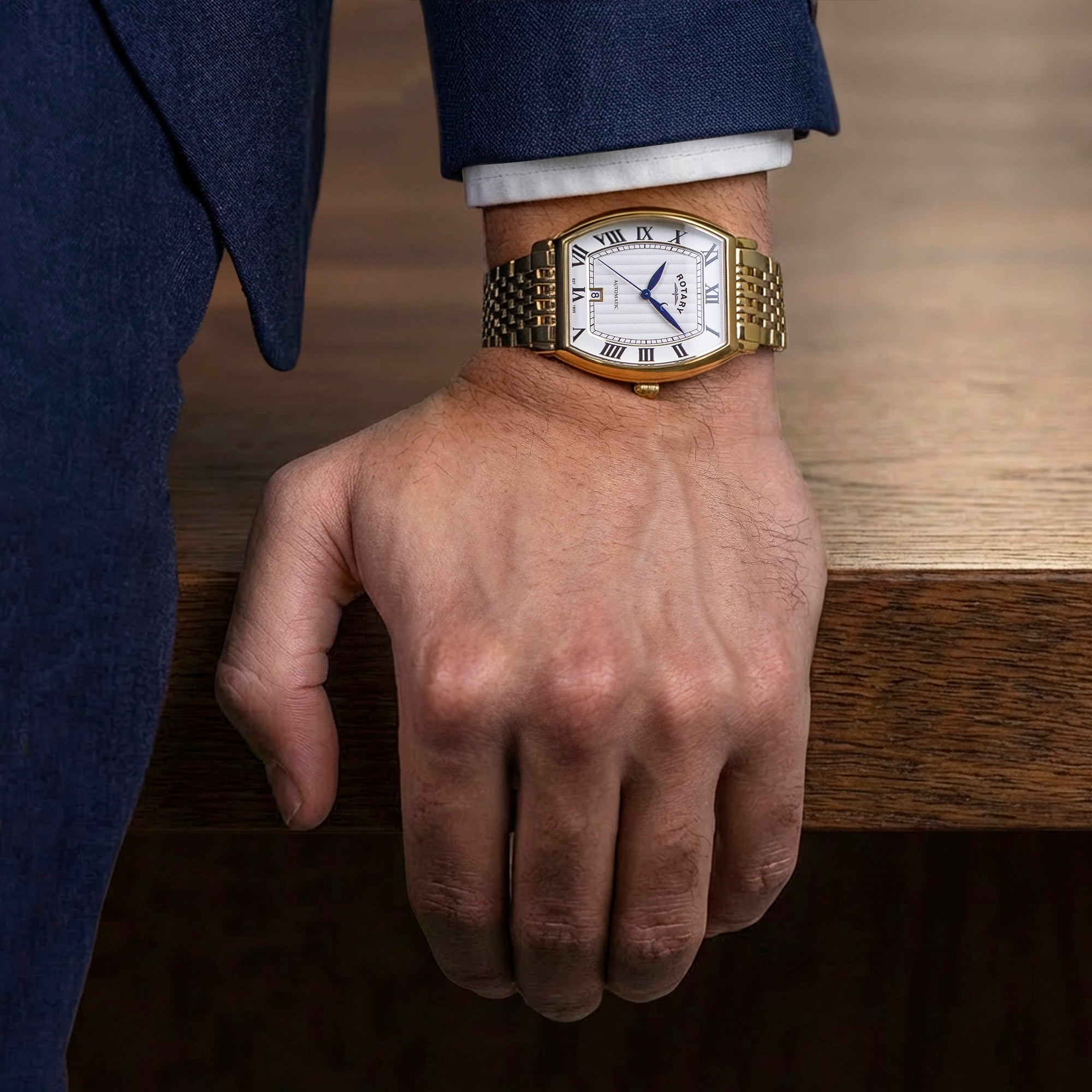Hand wearing a gold  rotary 1895 Tonneau automatic watch with a white face on a wooden surface
