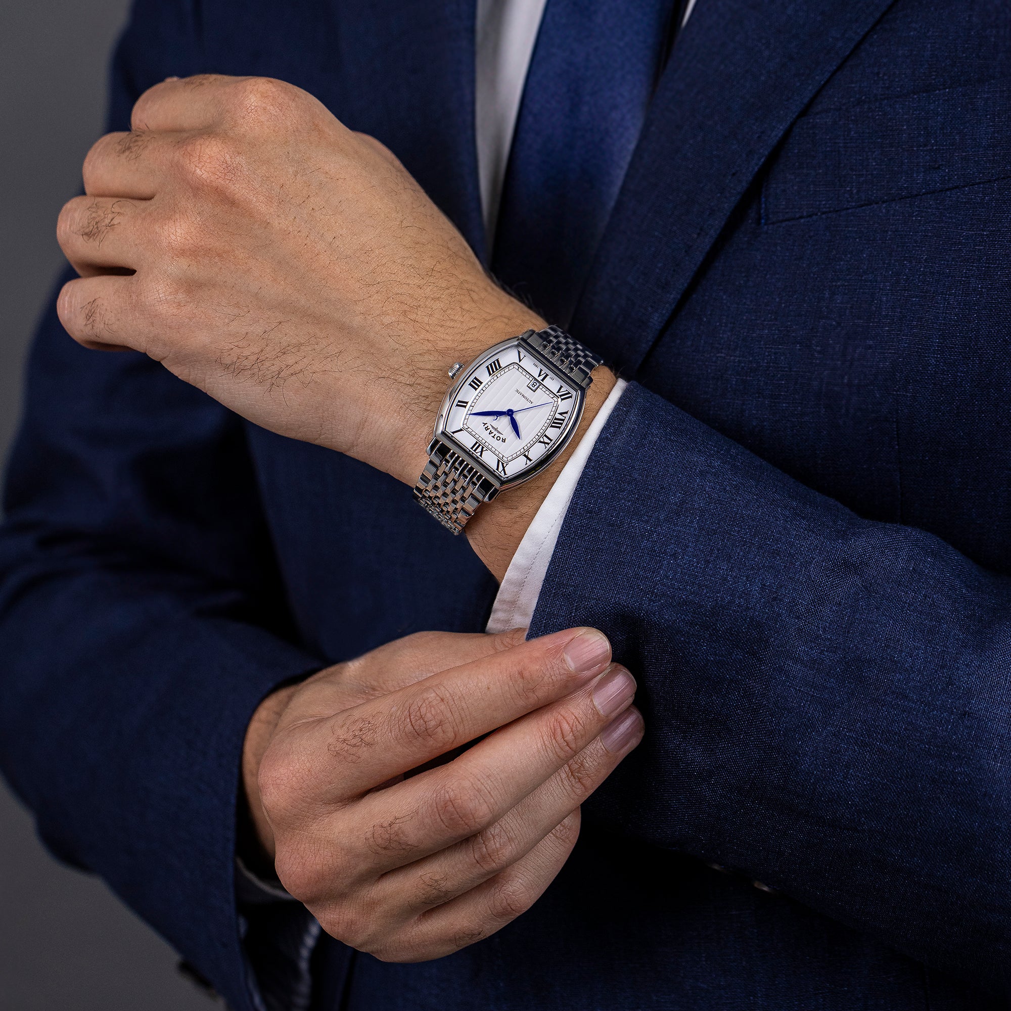 Man wearing a silver  rotary 1895 Tonneau automatic watch on a dark suit jacket against a gray background